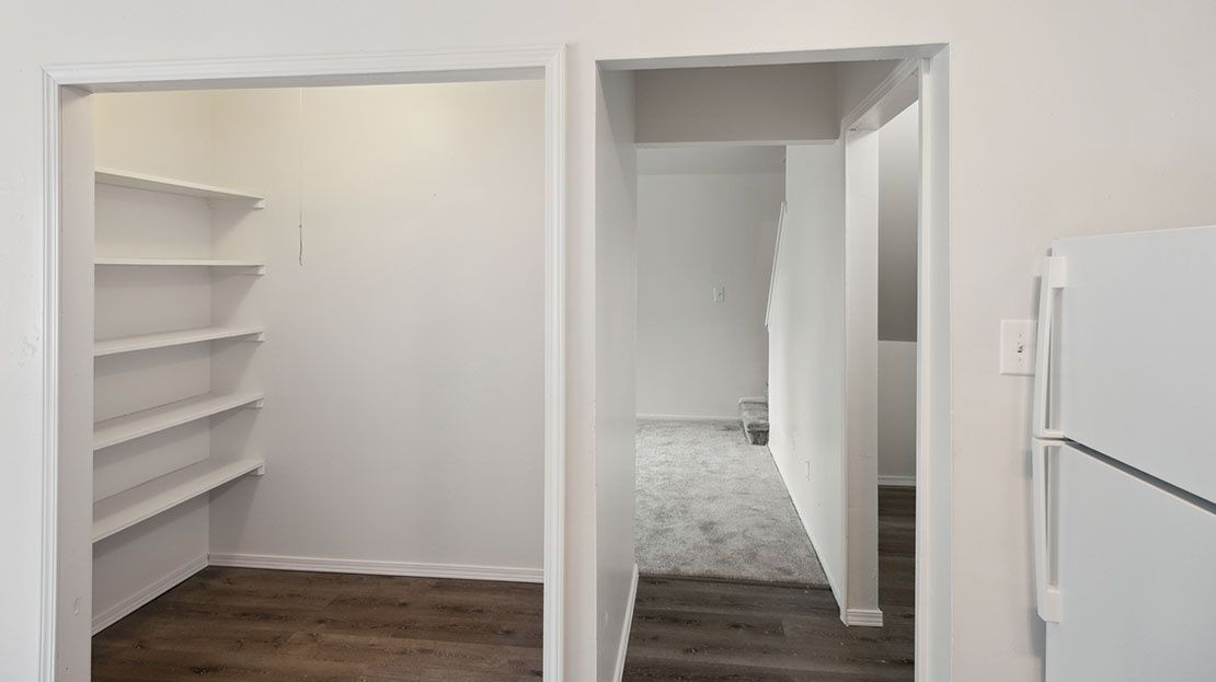 Empty white walk-in closet with built-in shelves, a doorway, and a white refrigerator at right.
