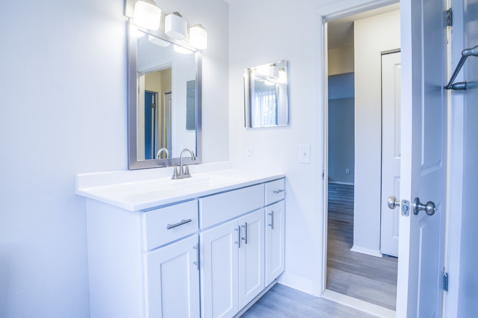 Bright white bathroom vanity with mirror and wall lights, open door to hallway and closet.