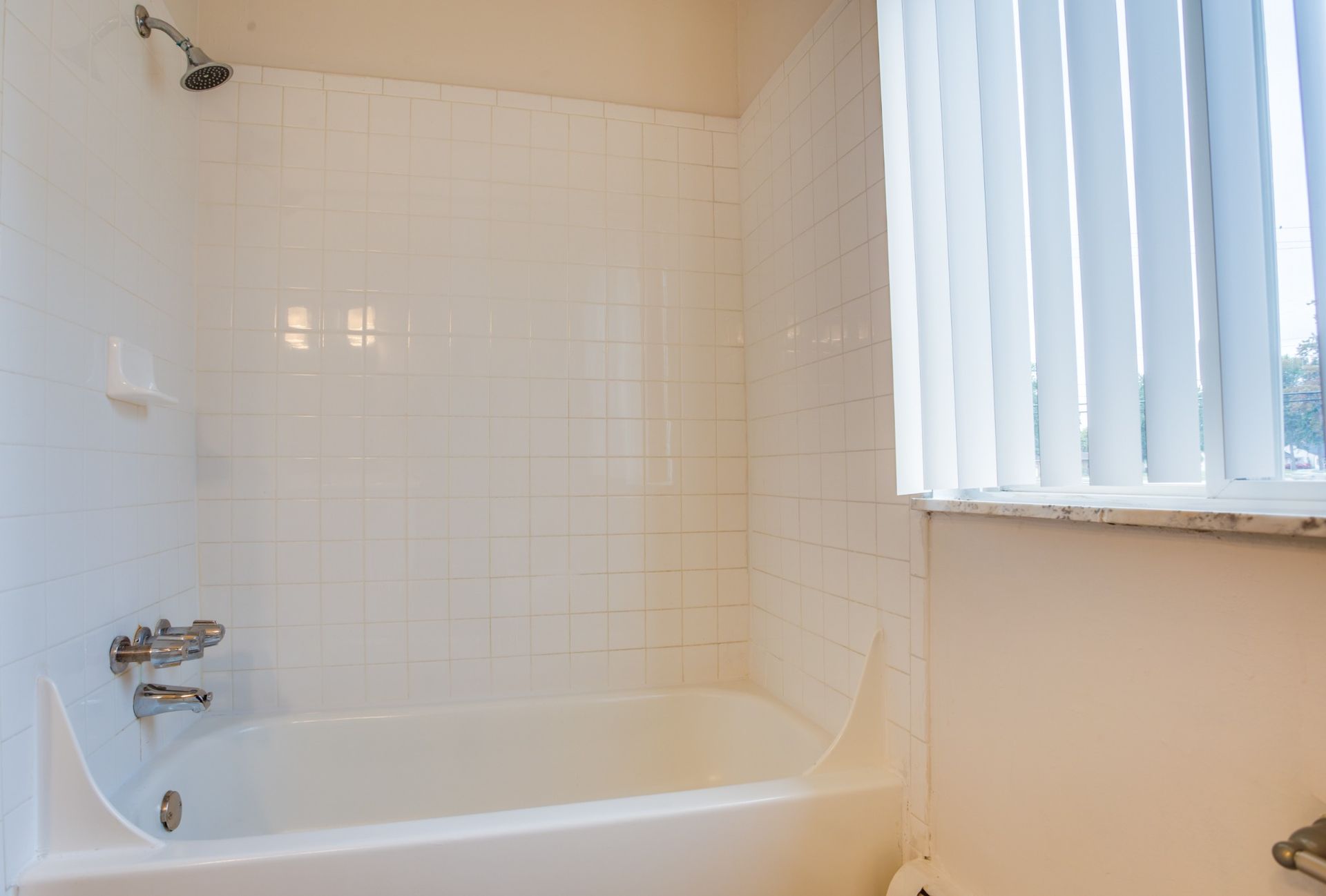 White tiled bathroom with a bathtub, shower fixtures, and a window with vertical blinds.
