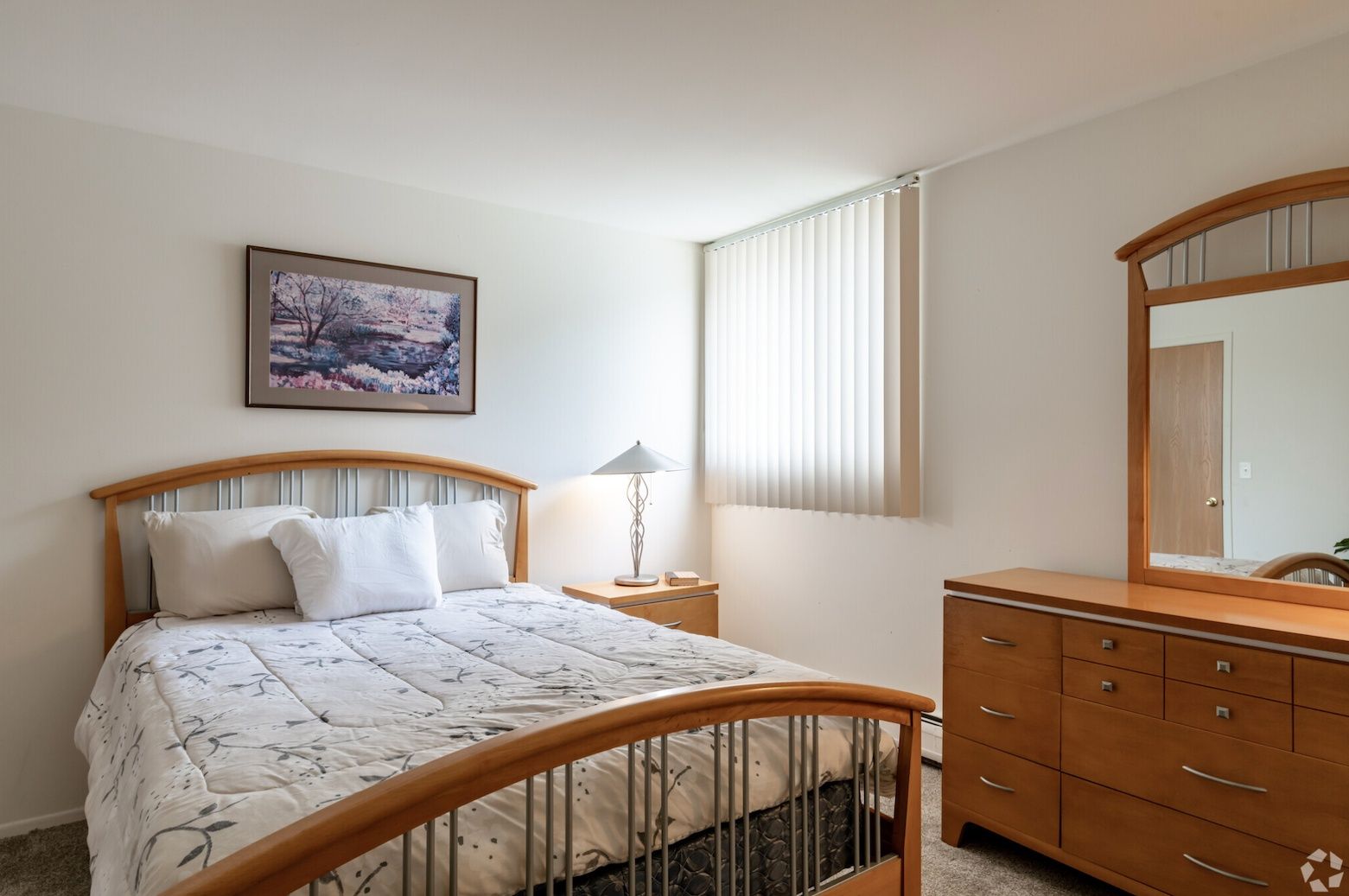 Bright bedroom with wooden bed, dresser, lamp, mirror, and window blinds
