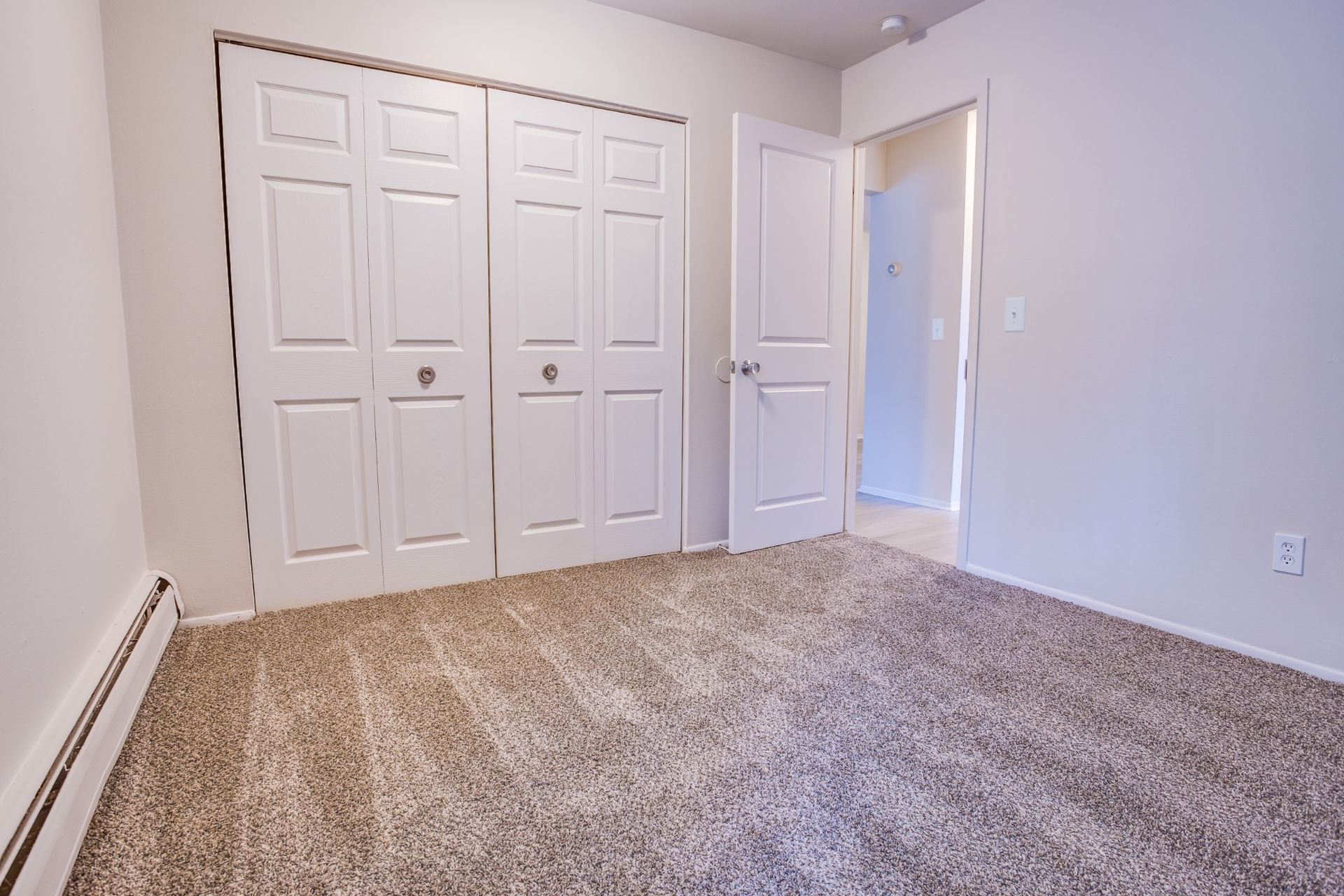 Empty carpeted bedroom with white double closet doors and an open door to a hallway