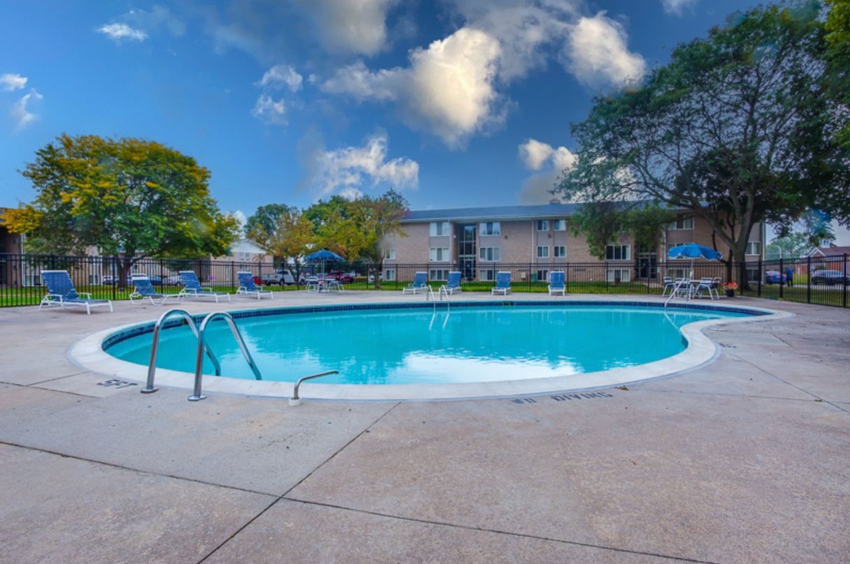 Outdoor apartment pool with blue water, white deck, and trees under a partly cloudy sky
