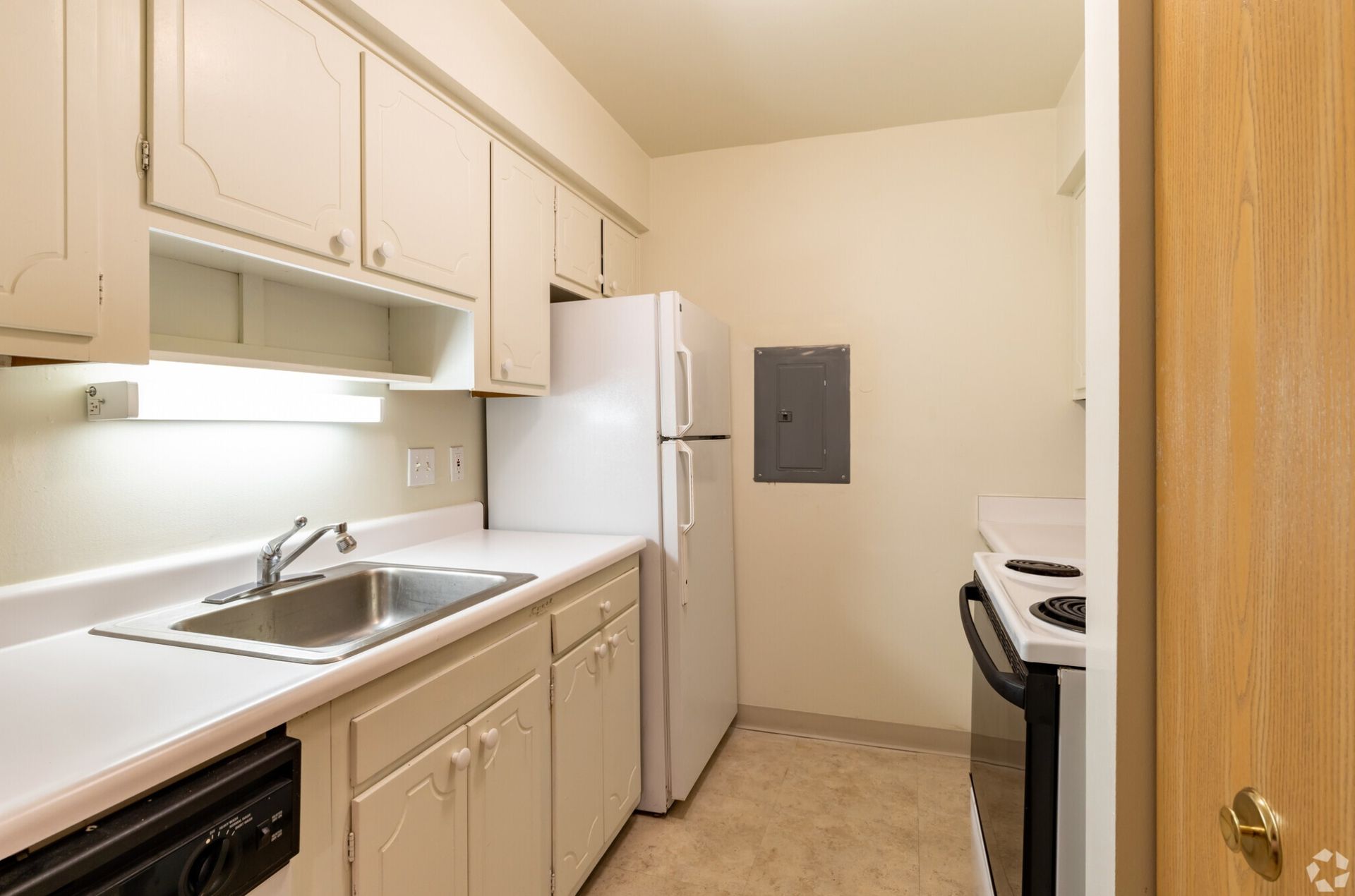 Small beige kitchen with white cabinets, sink, refrigerator, stove, and a wall electrical panel