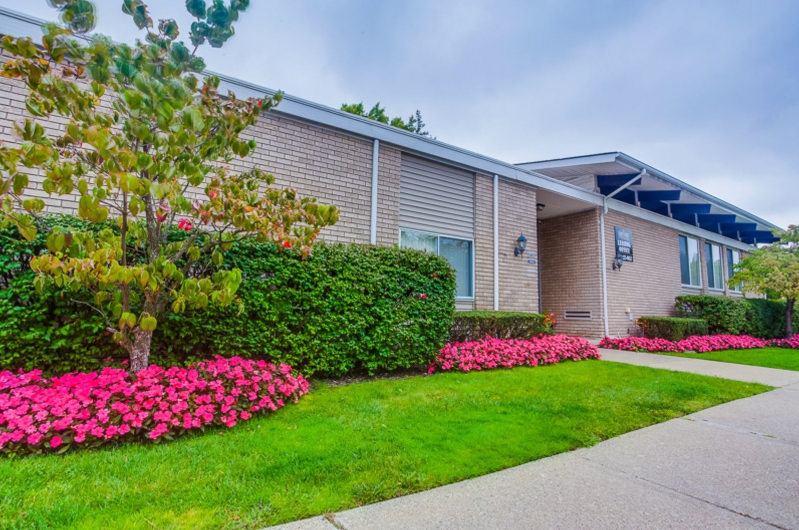 Suburban apartment building with pink flower beds, green lawn, and sidewalk under cloudy sky