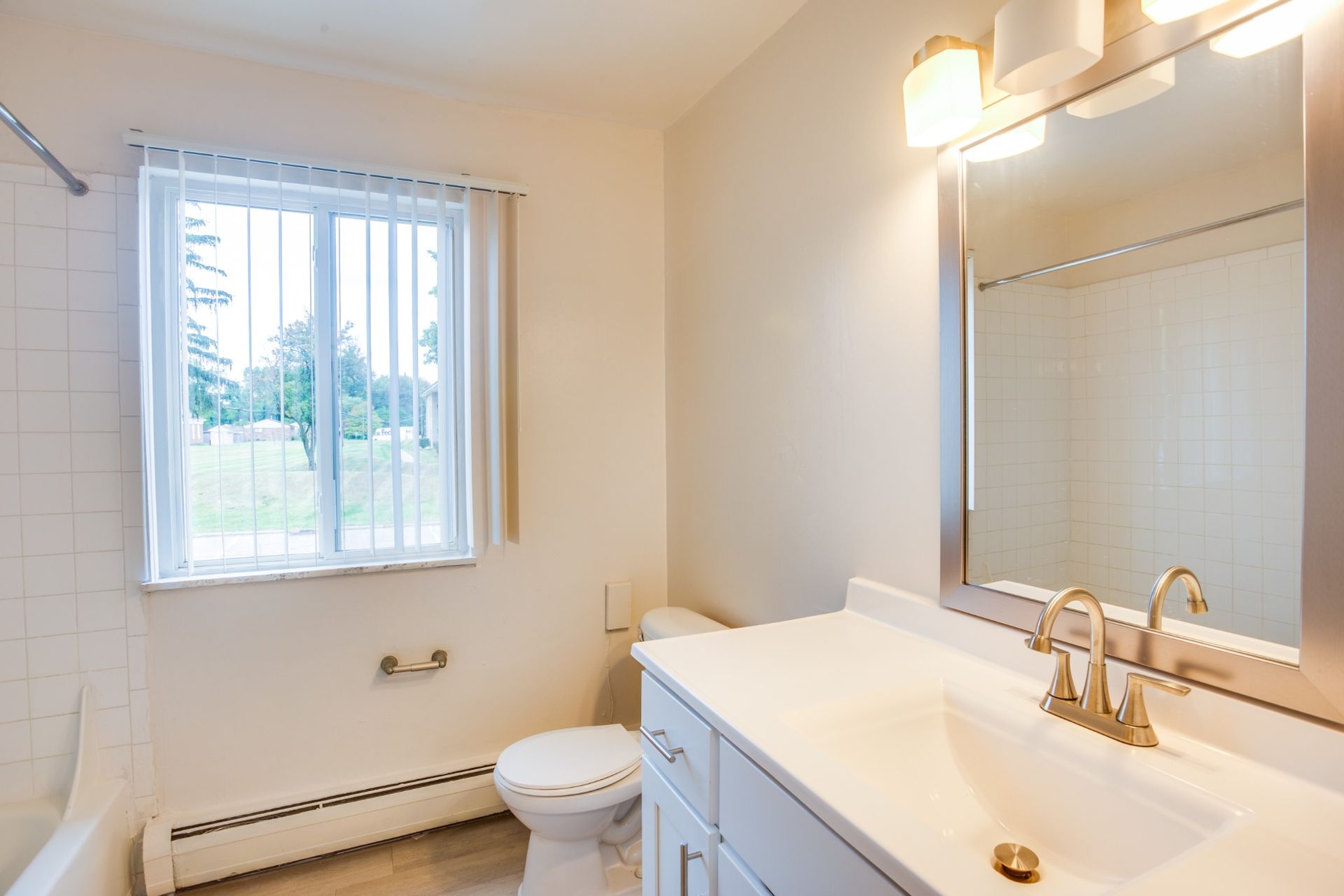 Bright bathroom with white vanity, gold faucet, large mirror, and a window with blinds.