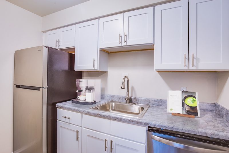 Small white kitchen with stainless fridge, sink, cabinets, and marble-pattern countertop.