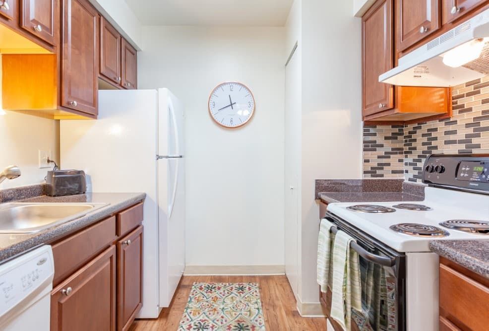 Narrow galley kitchen with wood cabinets, white appliances, and a wall clock at the end.