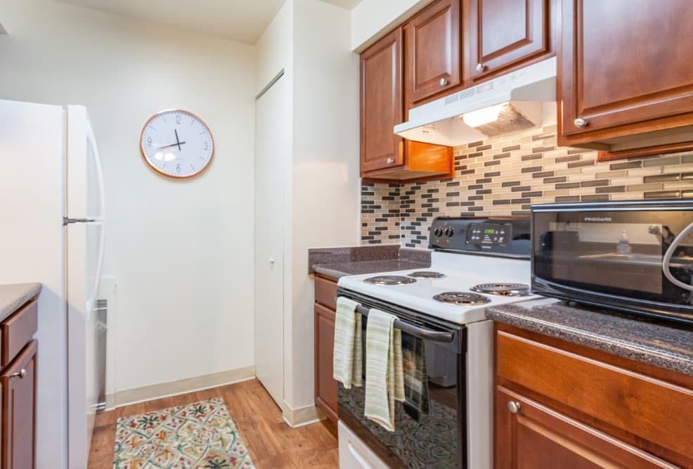 Compact kitchen with white appliances, wooden cabinets, mosaic backsplash, and a wall clock.