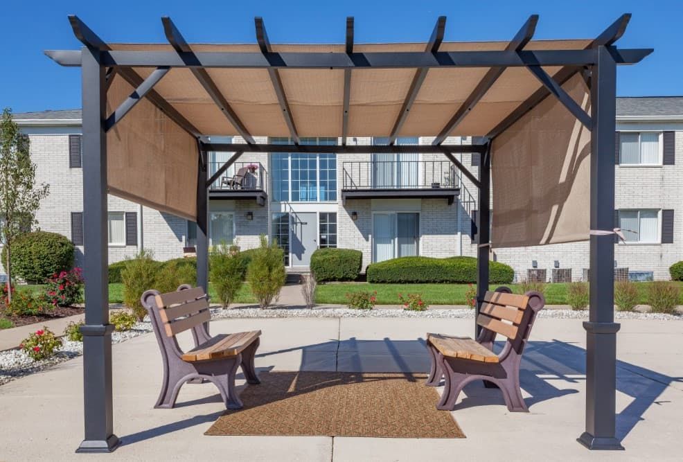 Outdoor pergola seating area with two wooden benches, tan canopy panels, and apartment buildings behind.