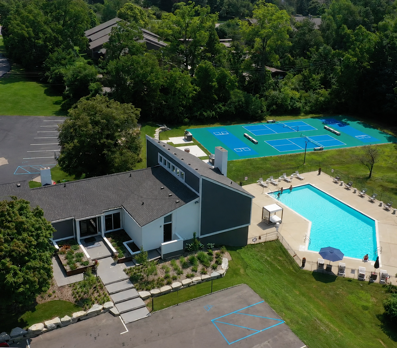 Aerial view of a poolside recreation area with a building and blue tennis courts surrounded by trees.