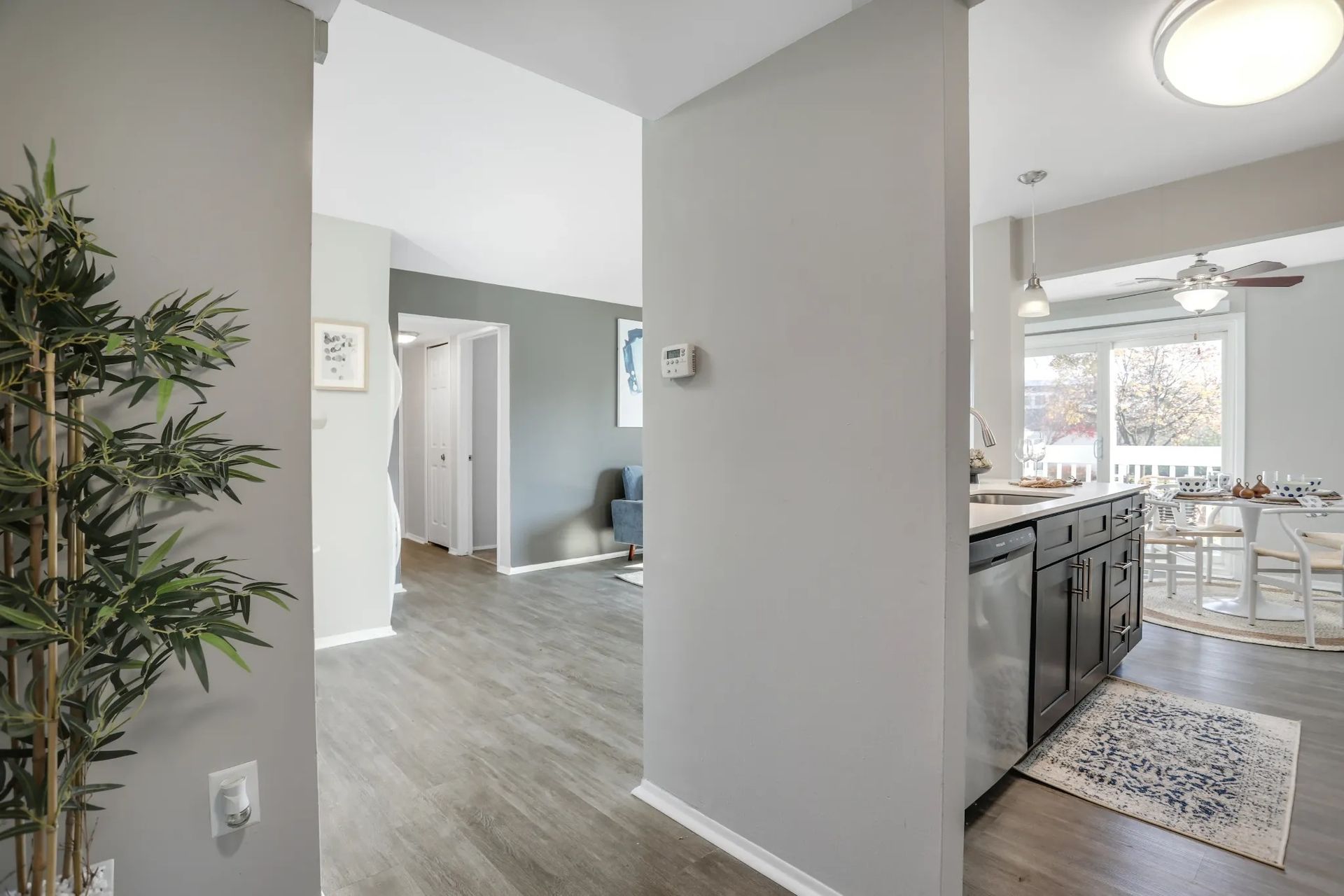Bright open hallway beside a kitchen with gray cabinets, light wood floors, and a windowed dining area.