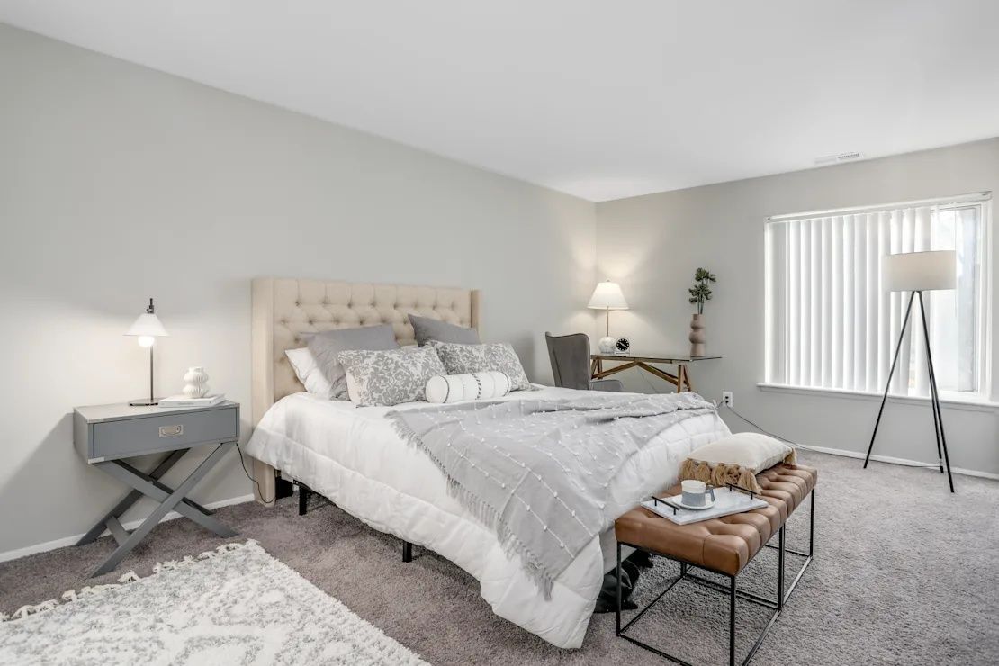 Bright bedroom with a tufted bed, gray decor, bedside lamp, bench, and window with blinds.