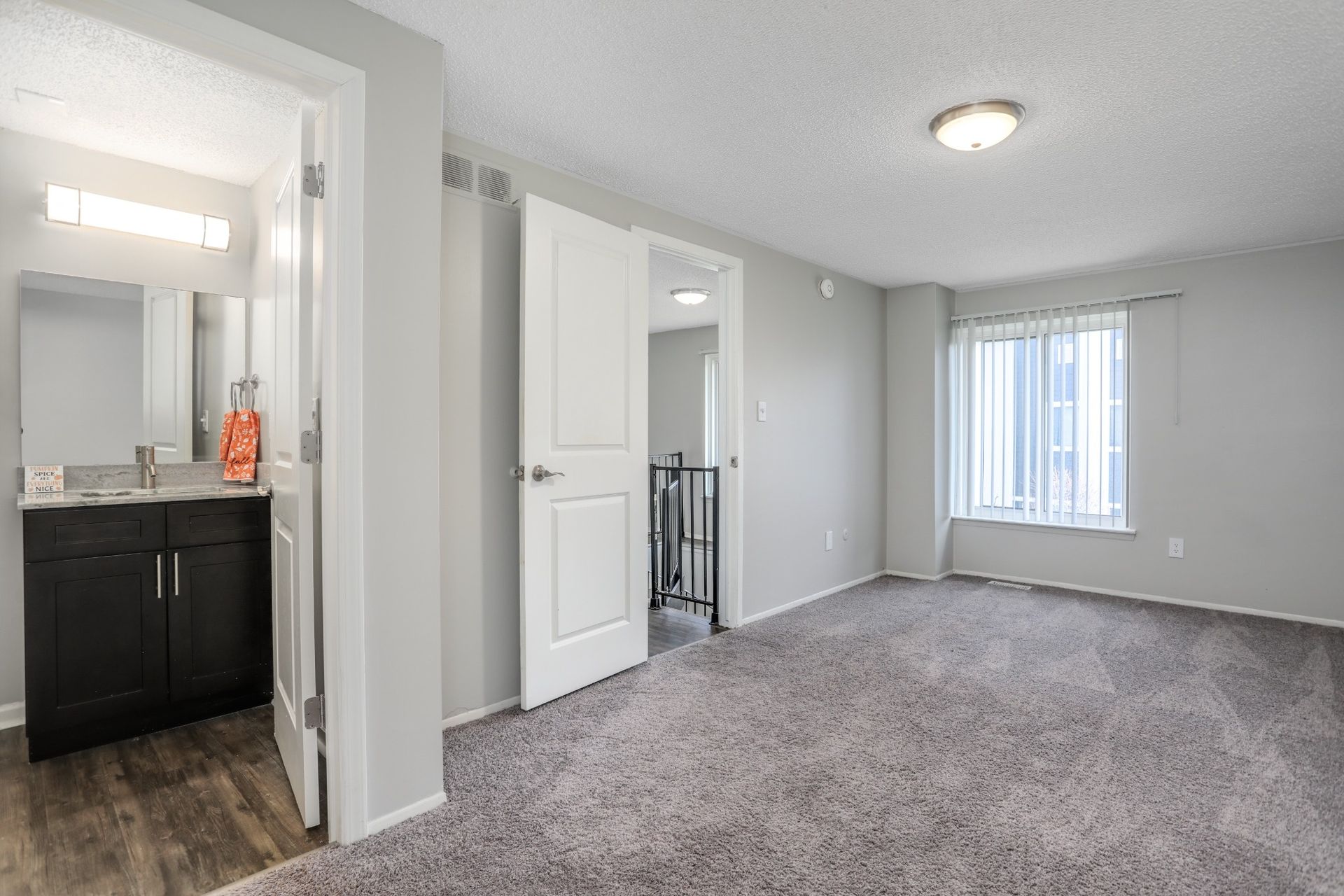 Empty gray-carpeted living room with white walls, a window, and a doorway to a bathroom.