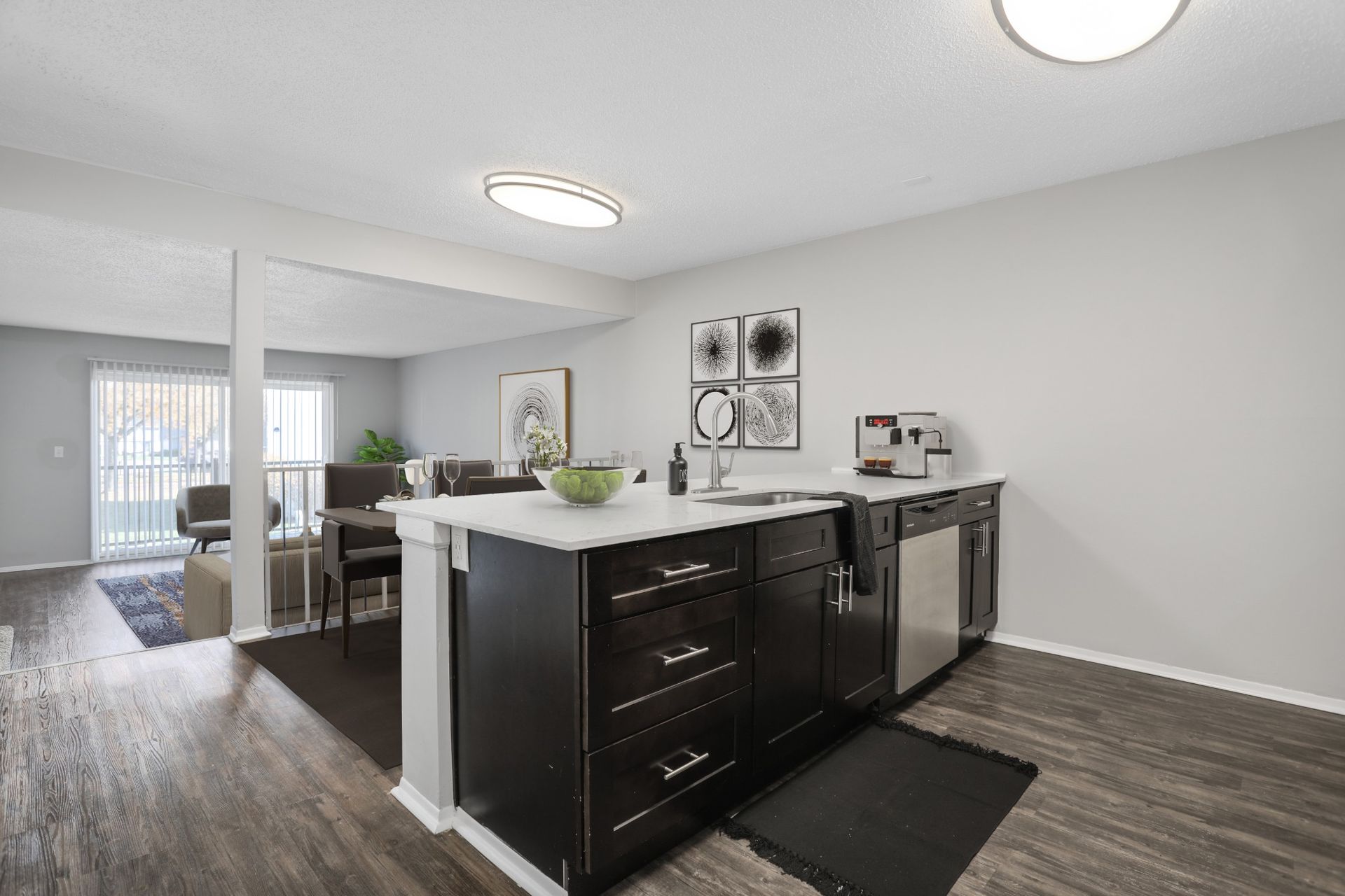Open-plan kitchen with dark island cabinets, white countertops, and gray flooring leading to a bright living area