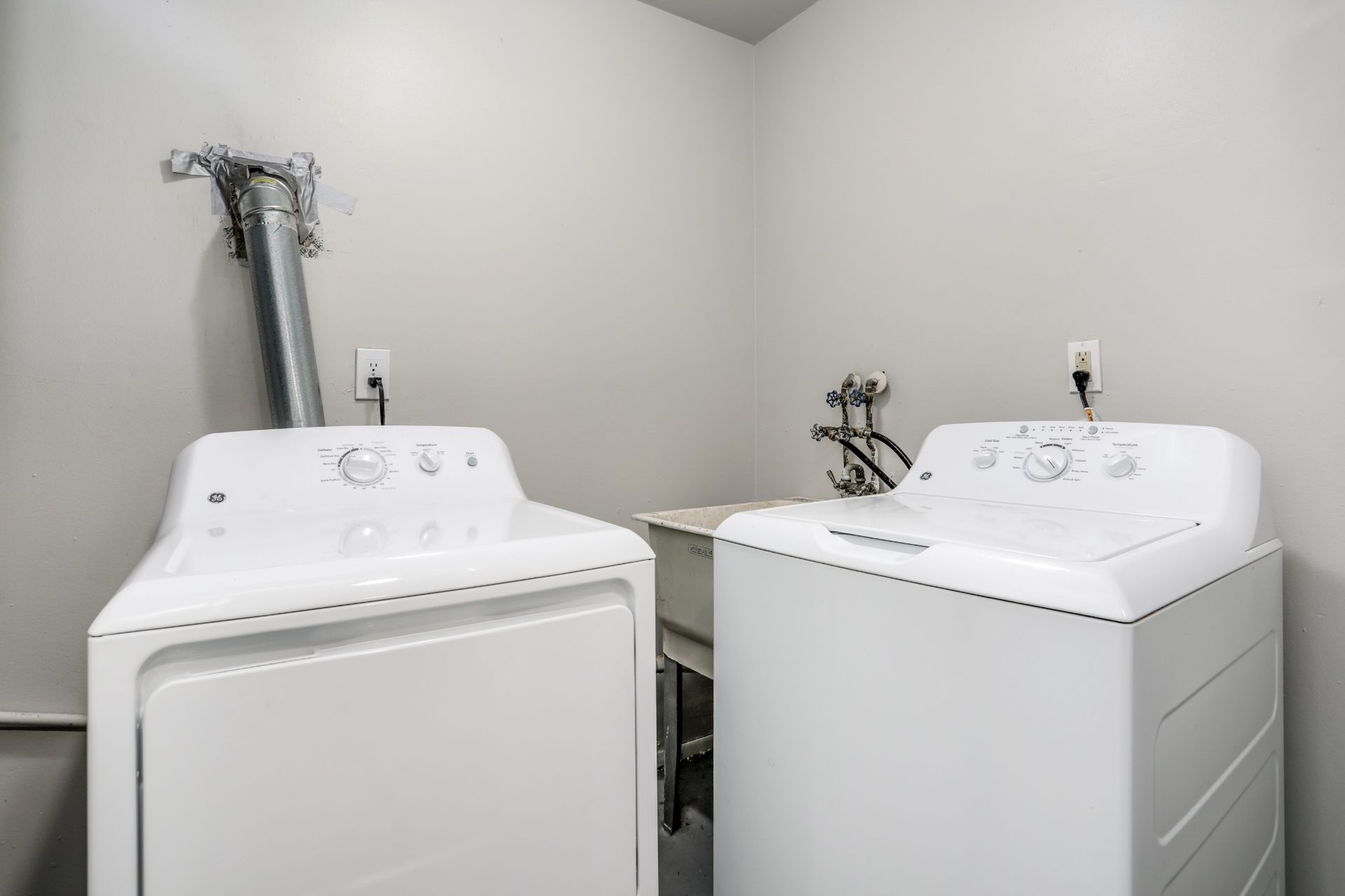 Laundry room with a white washer and dryer under utility hookups, in a plain gray corner.