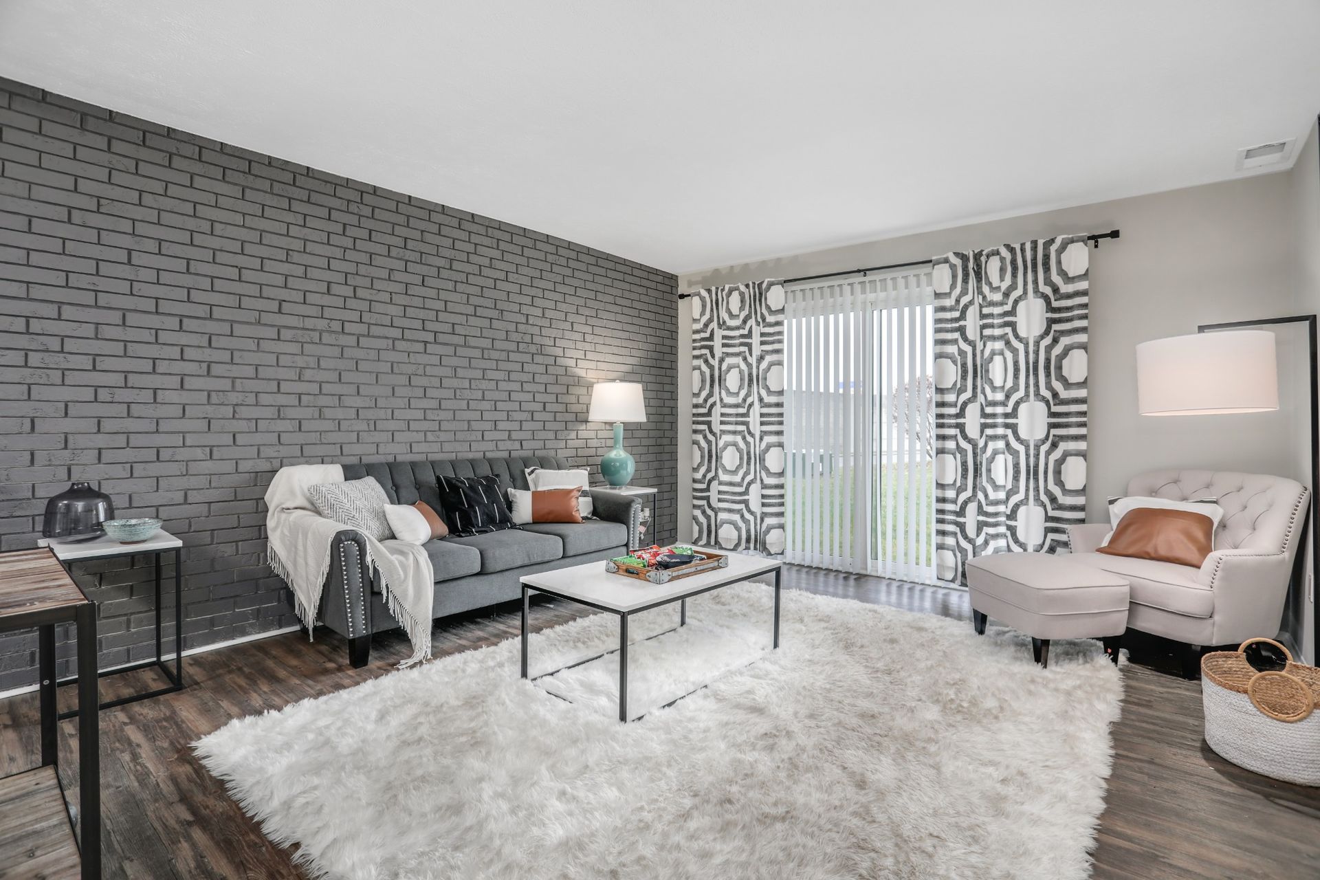 Modern living room with gray brick wall, white shag rug, glass coffee table, and patterned curtains.