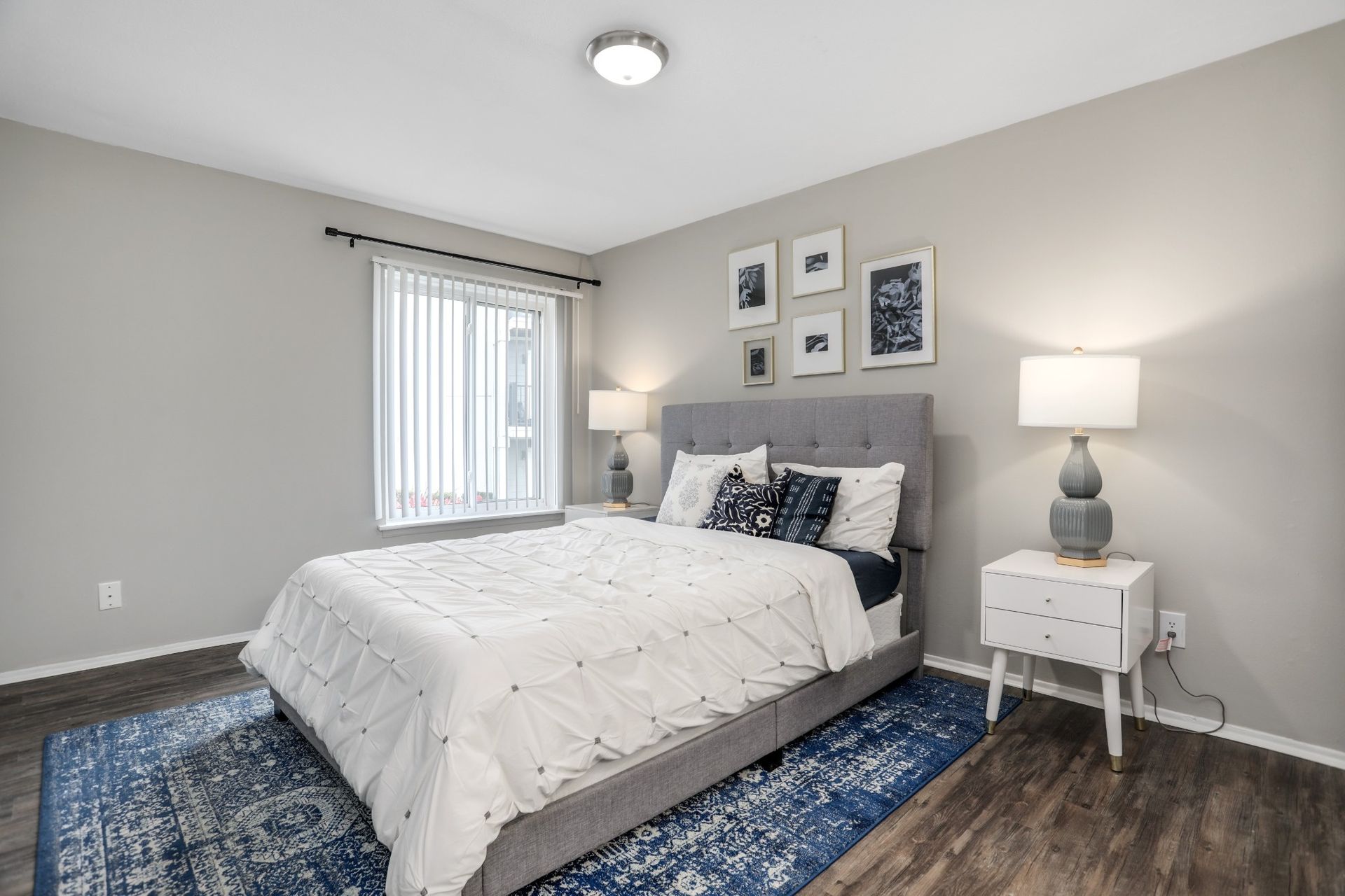 Bright bedroom with a gray bed, blue rug, white nightstand, and window with vertical blinds.