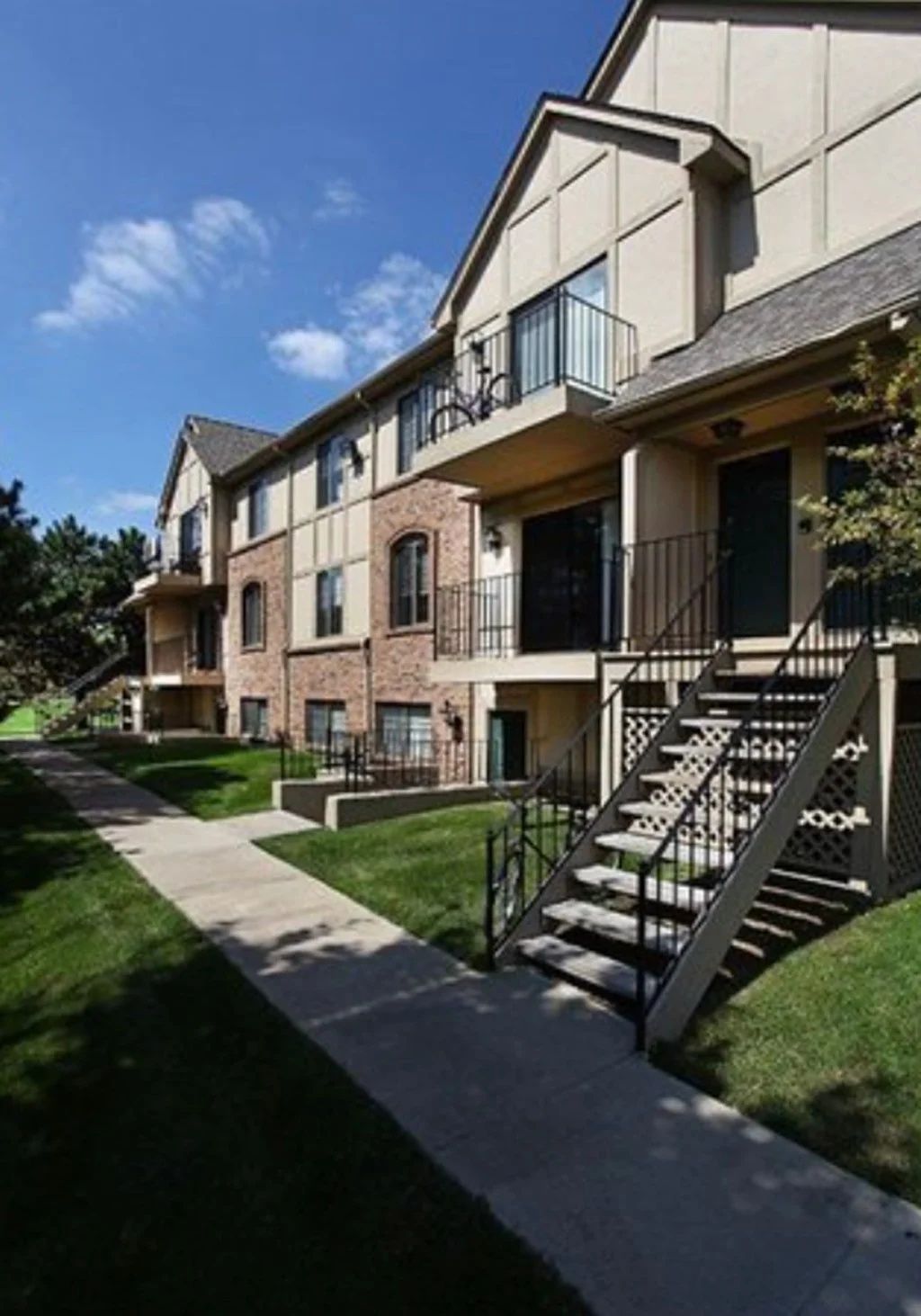 Apartment building exterior with stairs, balconies, and a concrete walkway beside a grassy lawn