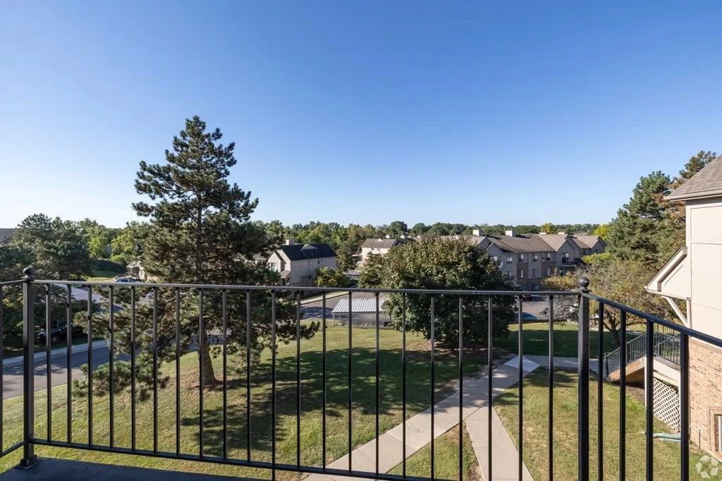 Balcony view of a green courtyard, trees, rooftops, and a clear blue sky behind a black metal railing