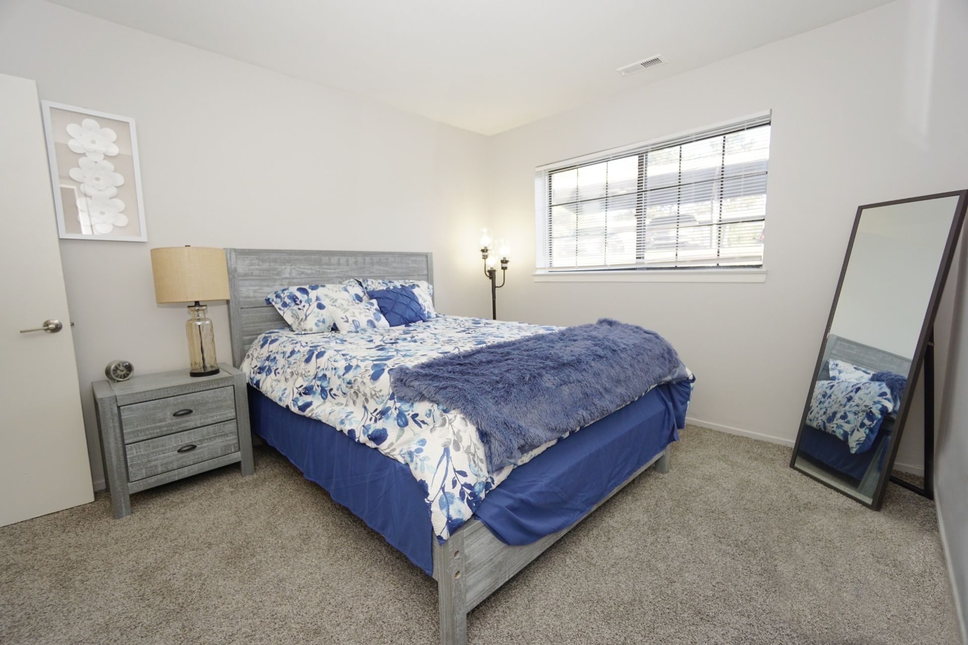 Bright bedroom with blue bedspread, nightstand lamp, window, and leaning floor mirror.