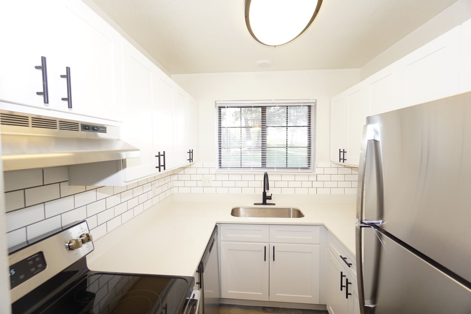 Bright white galley kitchen with stainless fridge, stove, sink under window, and black countertops