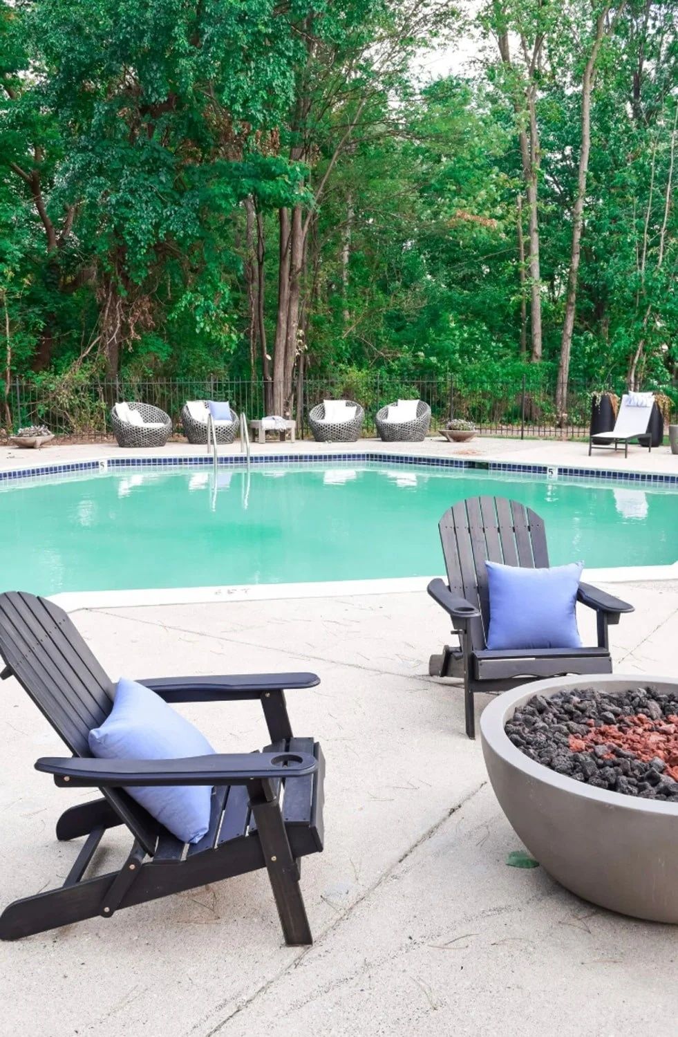 Backyard pool with two Adirondack chairs, blue pillows, and a fire pit surrounded by trees