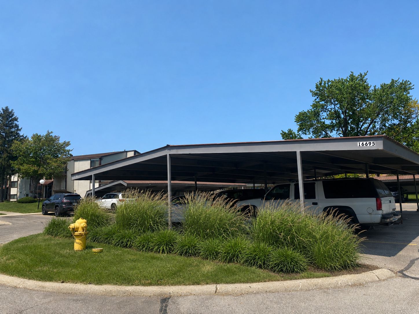 Covered parking lot beside a low apartment building, with cars, shrubs, and a yellow fire hydrant.