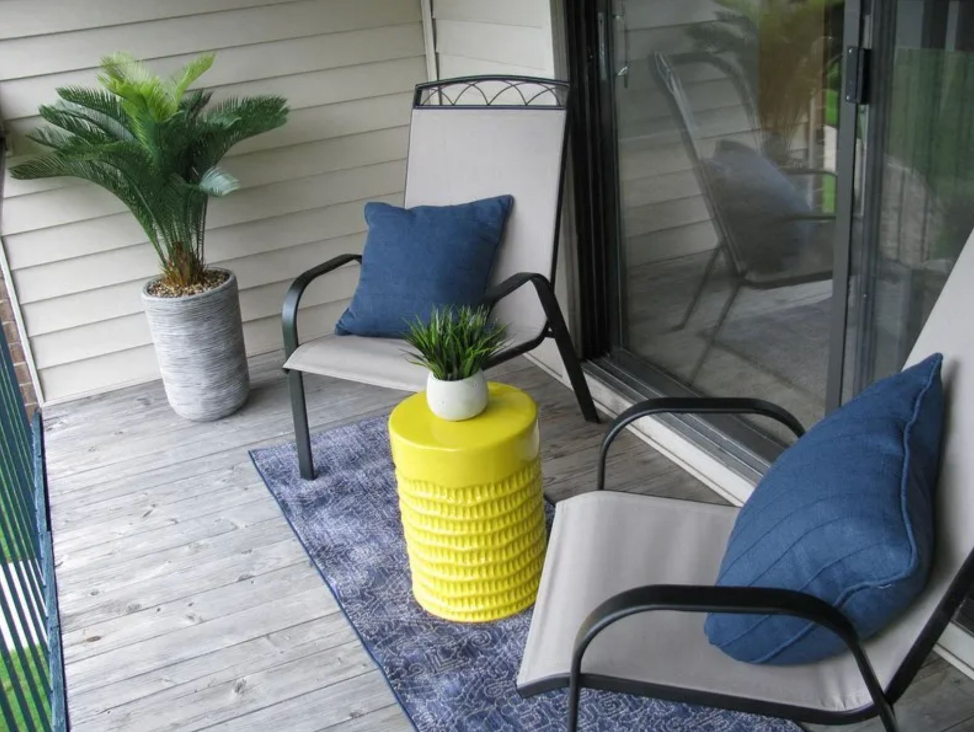 Patio seating area with two gray chairs, blue cushions, yellow side table, and potted plants on a wooden deck