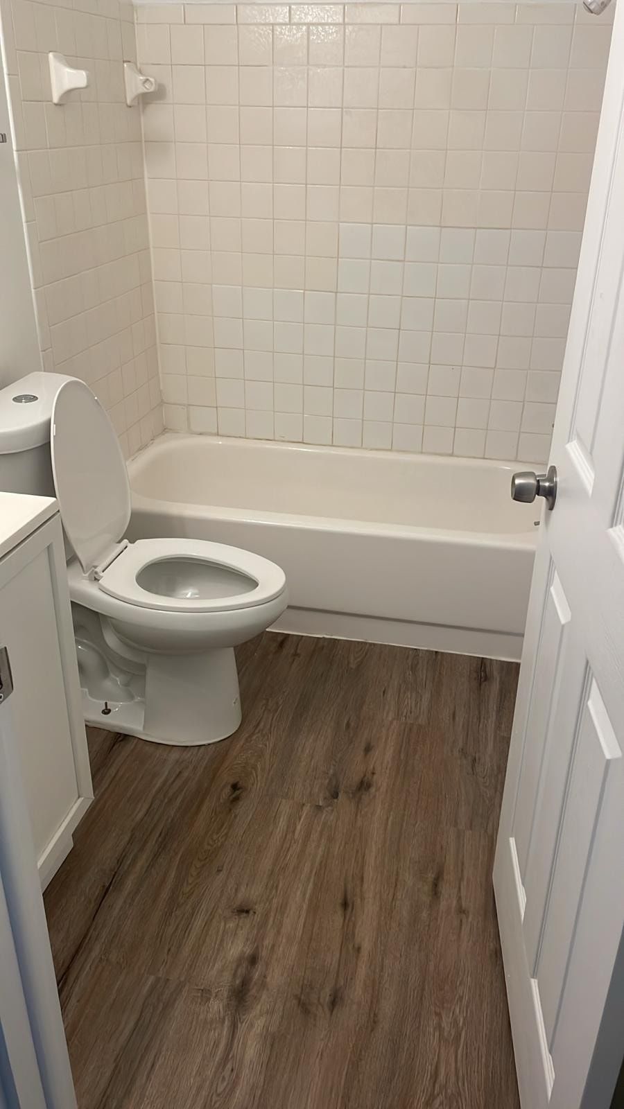 Small bathroom with toilet, bathtub, and wood-look floor; white walls and door open inward.