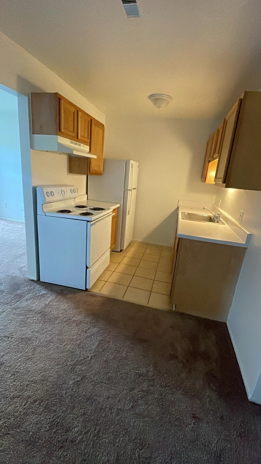 Small empty kitchen with white appliances, wood cabinets, and tiled floor beside carpeted living area