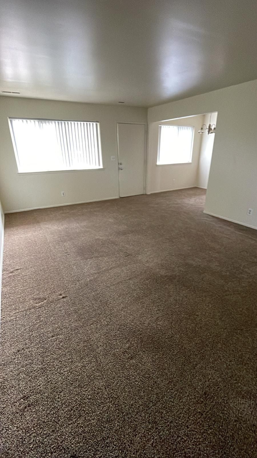 Empty carpeted living room with white walls, window blinds, and an open doorway to another room