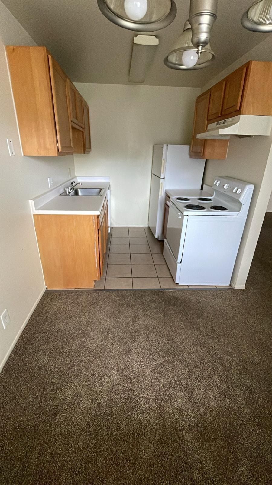 Small empty kitchen with tan cabinets, white stove and fridge, sink, and carpeted floor.