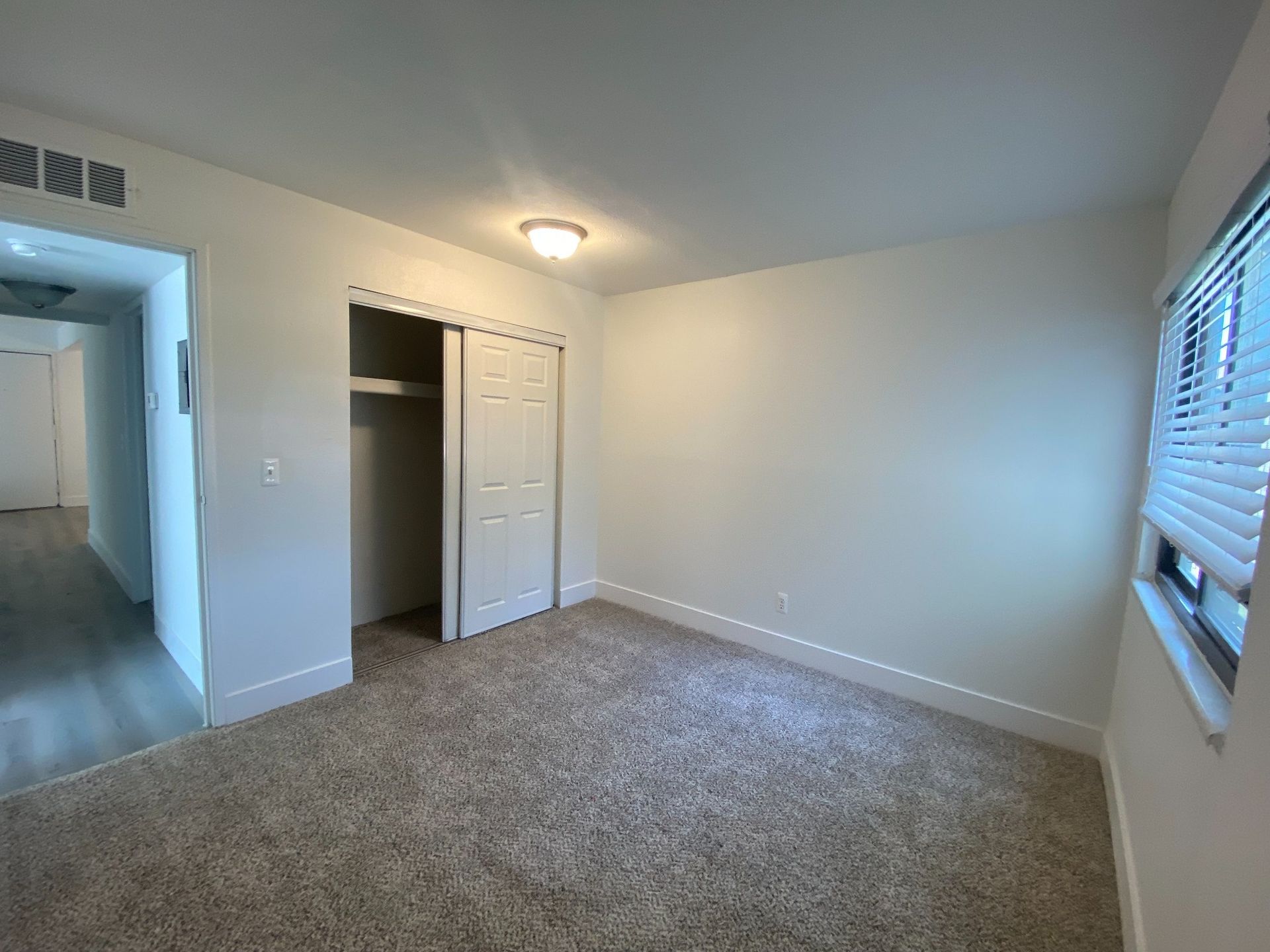 Empty carpeted bedroom with white walls, closet, and a window with blinds.