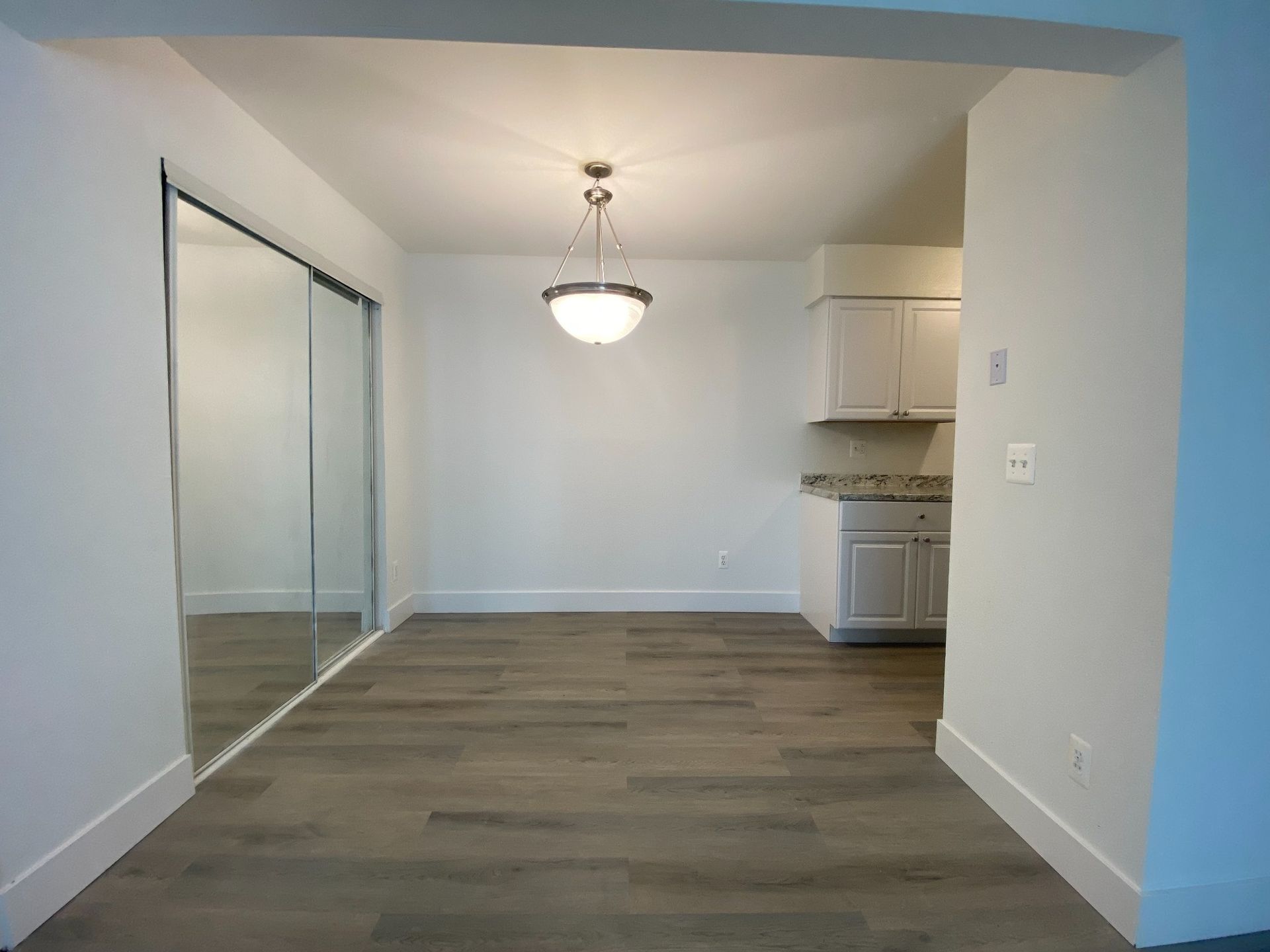 Empty dining area with wood floors, mirrored closet, and small kitchen nook in the back.