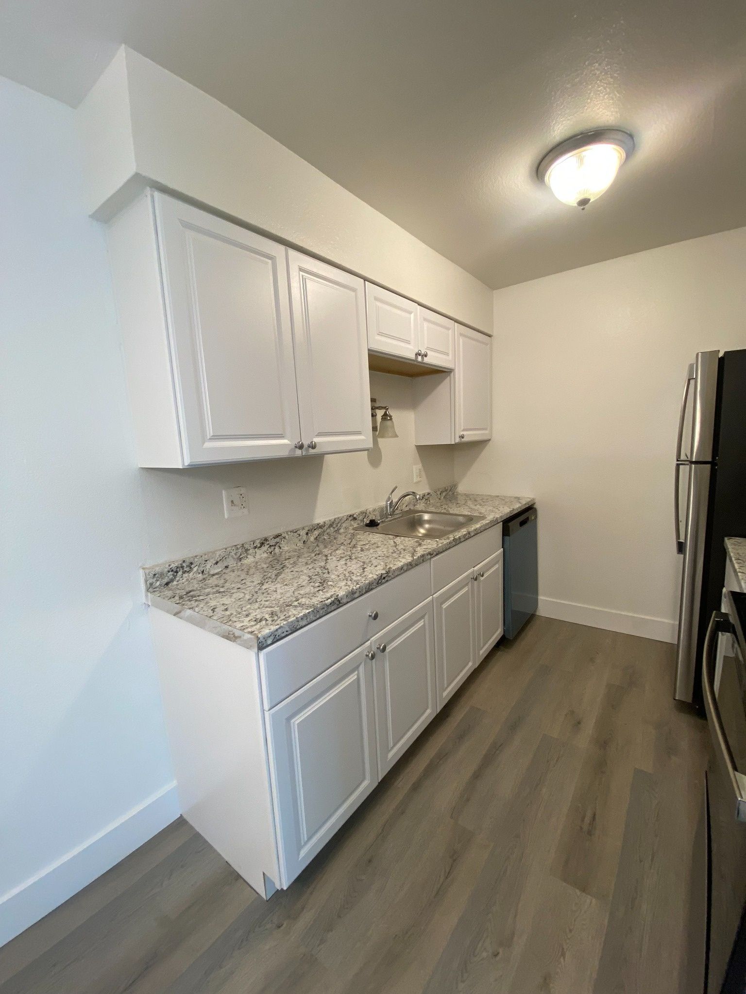 Empty modern kitchen with white cabinets, speckled countertop, wood-look floor, and stainless steel refrigerator