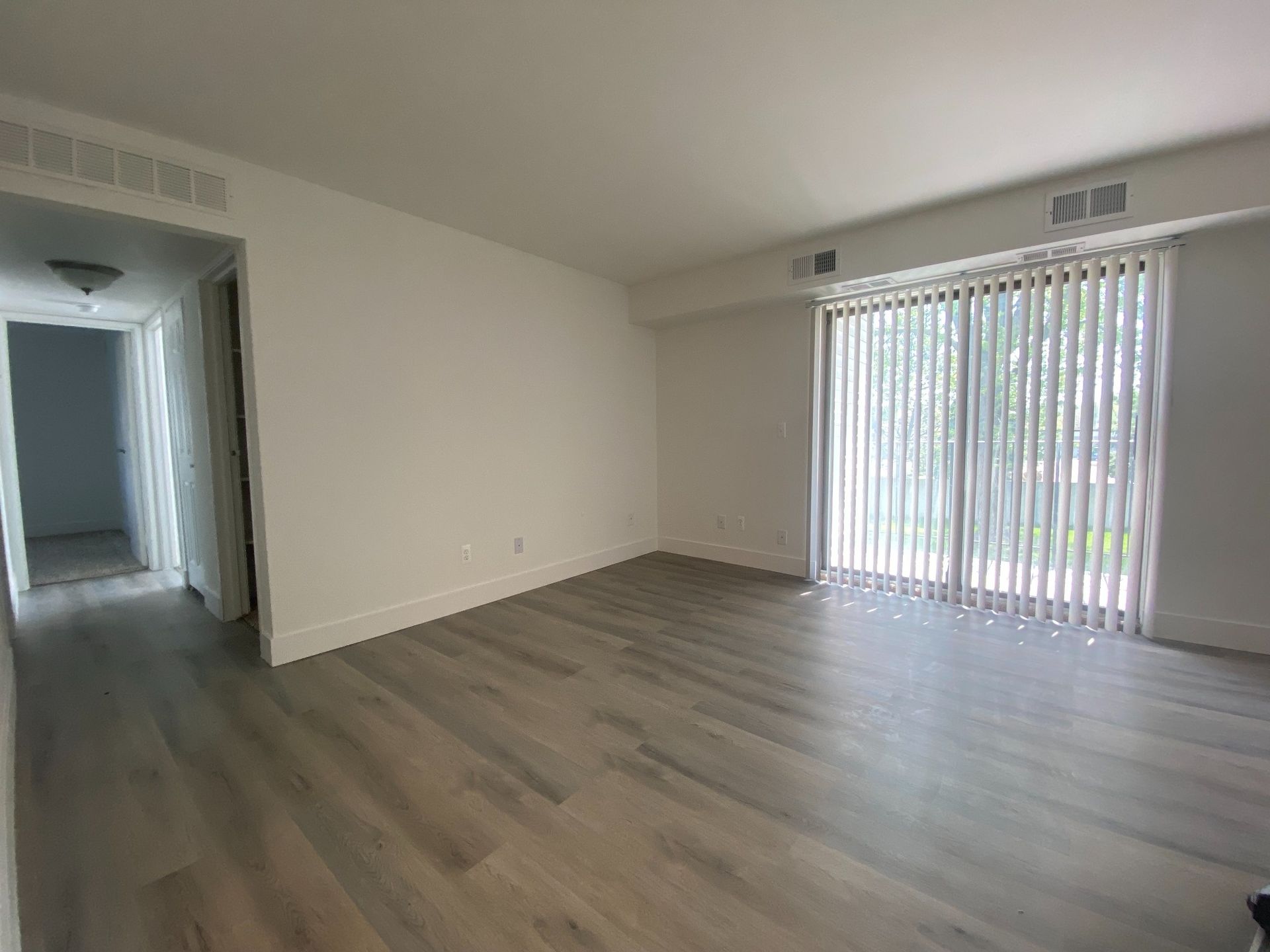 Empty modern living room with gray wood flooring, white walls, and sliding glass doors with vertical blinds