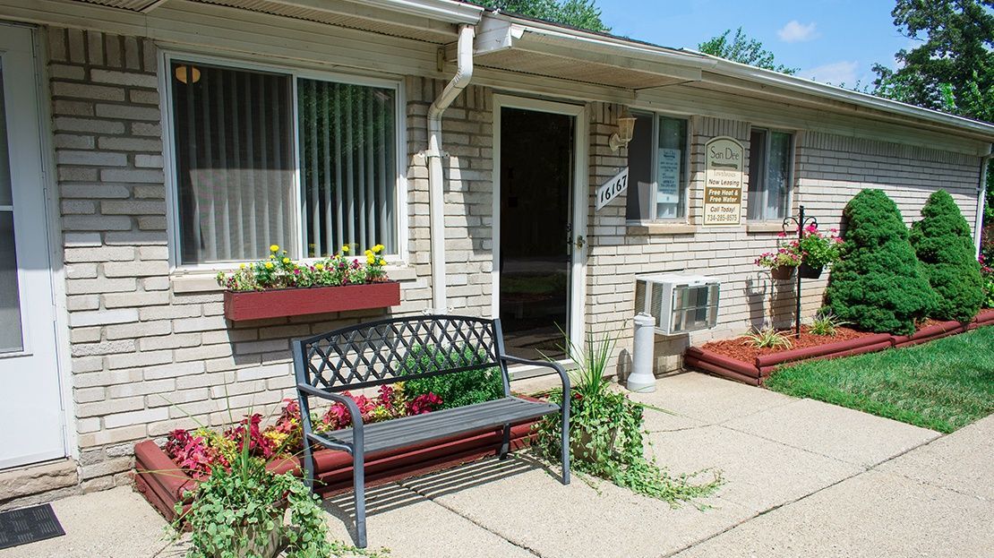 Front porch of a brick house with flower boxes, a bench, potted plants, and a concrete walkway.