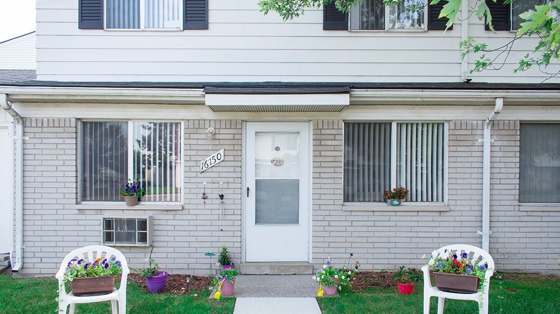 Front entrance of a gray house with white door, barred windows, flower beds, and potted plants by the walkway