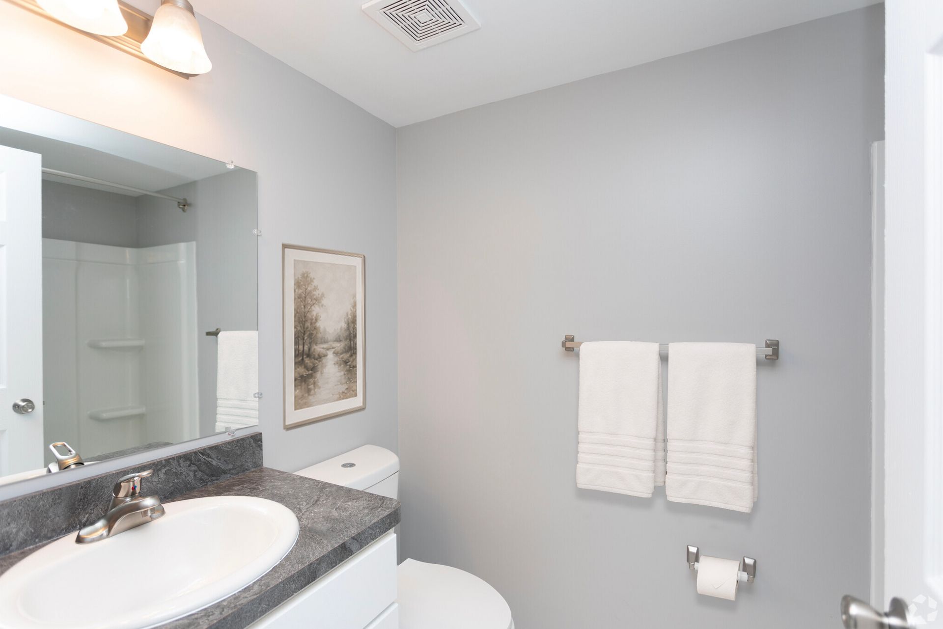 Bright bathroom with gray walls, granite vanity, mirror, and white towels by the toilet.