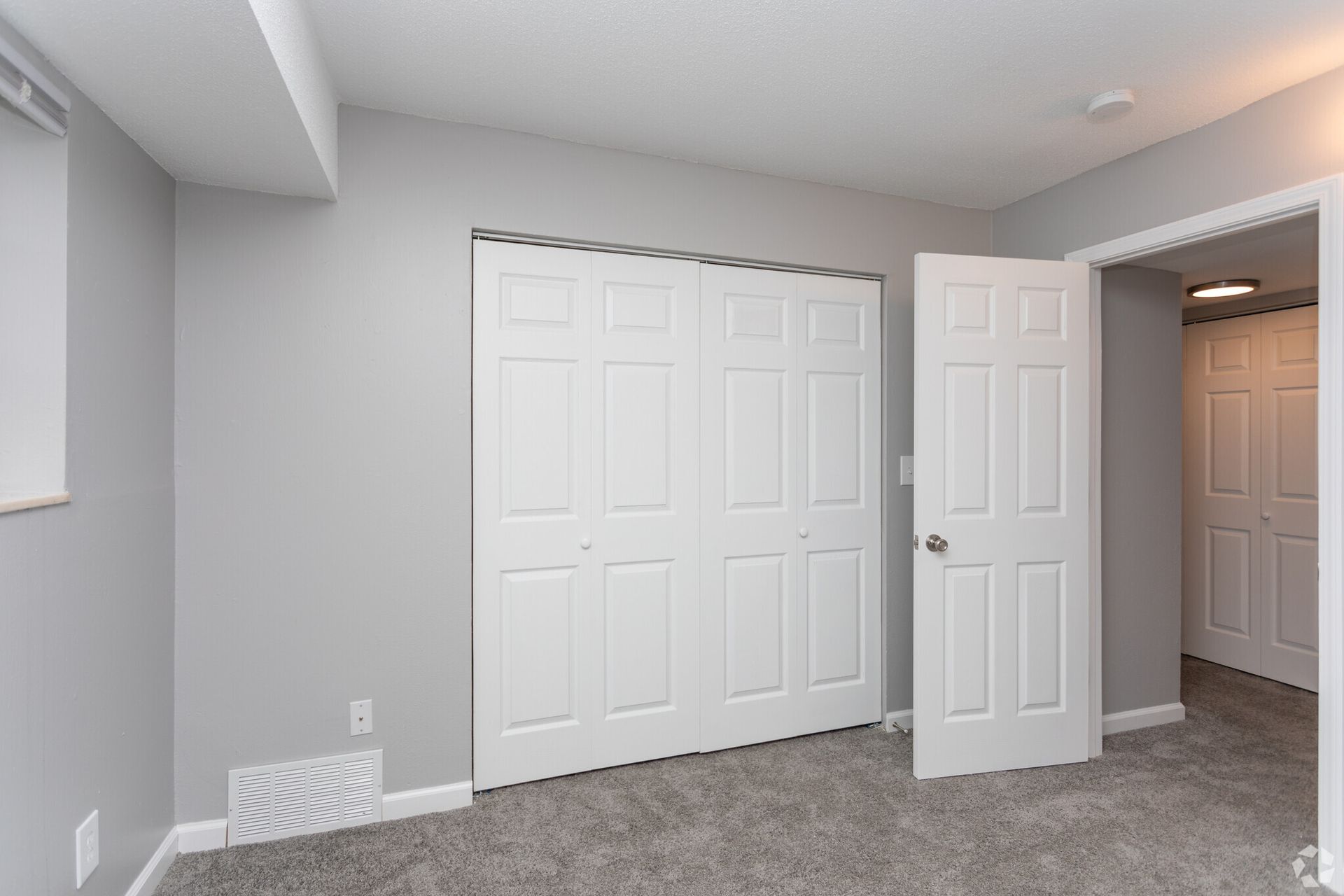 Empty carpeted bedroom with gray walls, white closet doors, and an open door to a hallway