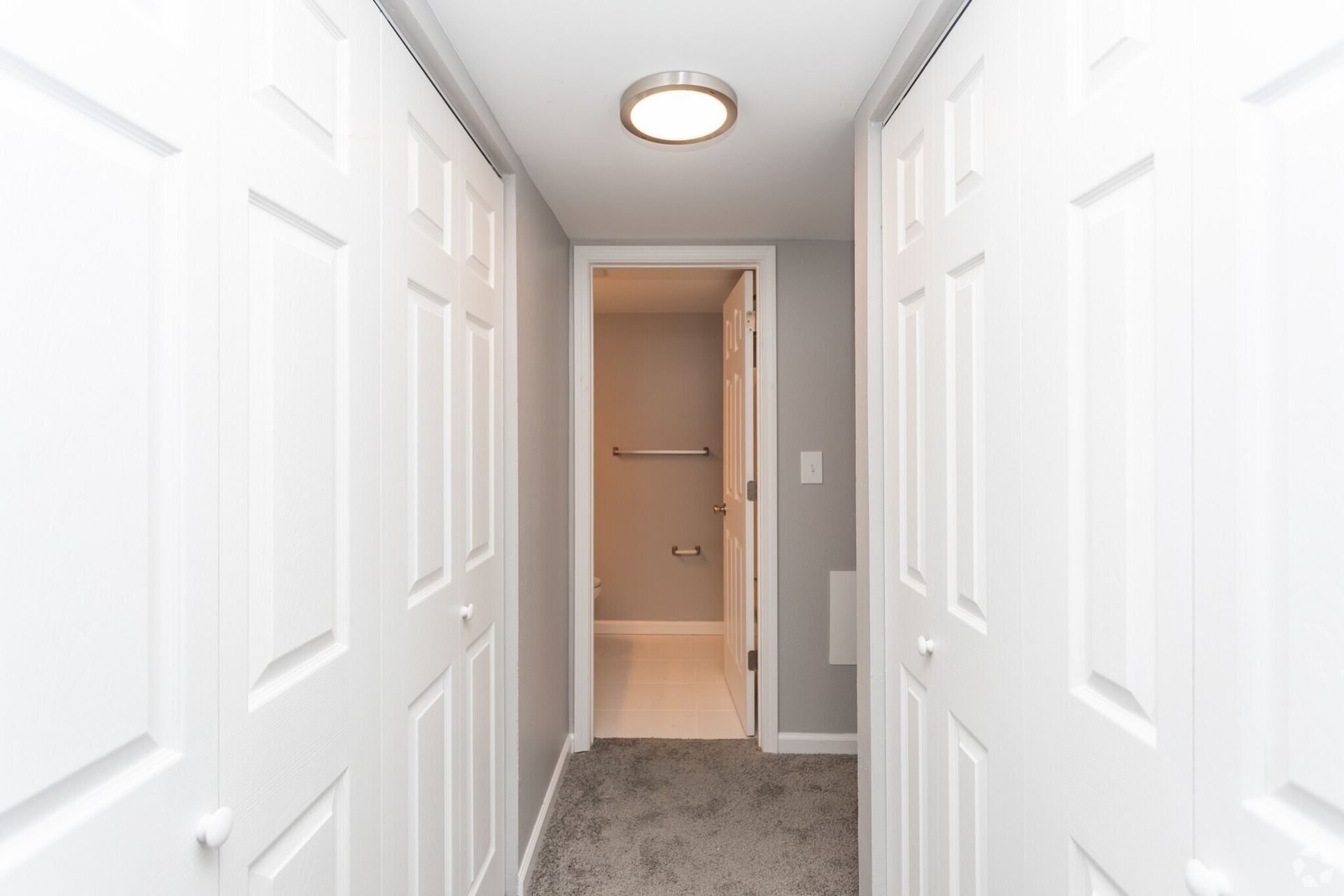 Narrow white hallway with mirrored closet doors, carpet, and an open doorway to a lit bathroom.