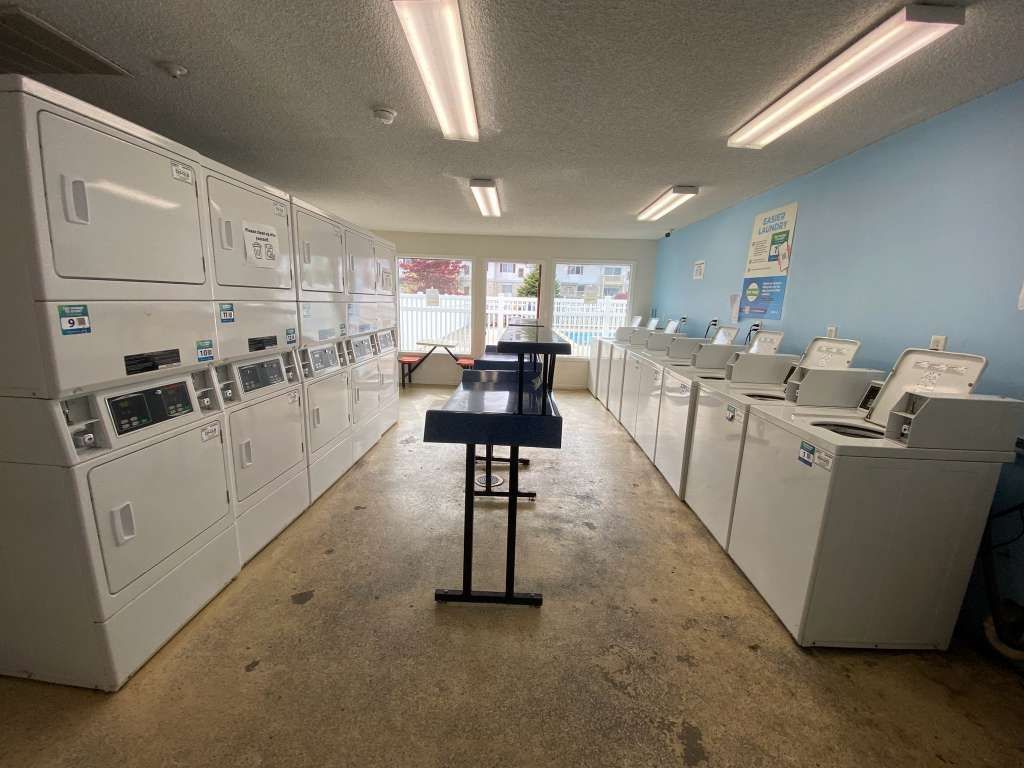 Laundromat interior with rows of white washing machines and dryers, and a folding table in the center.