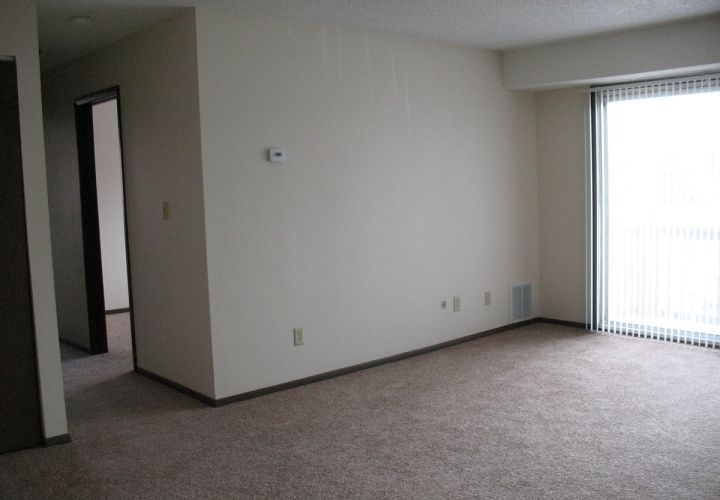 Empty beige living room with carpet, doorway on left, and sliding glass door with vertical blinds on the right