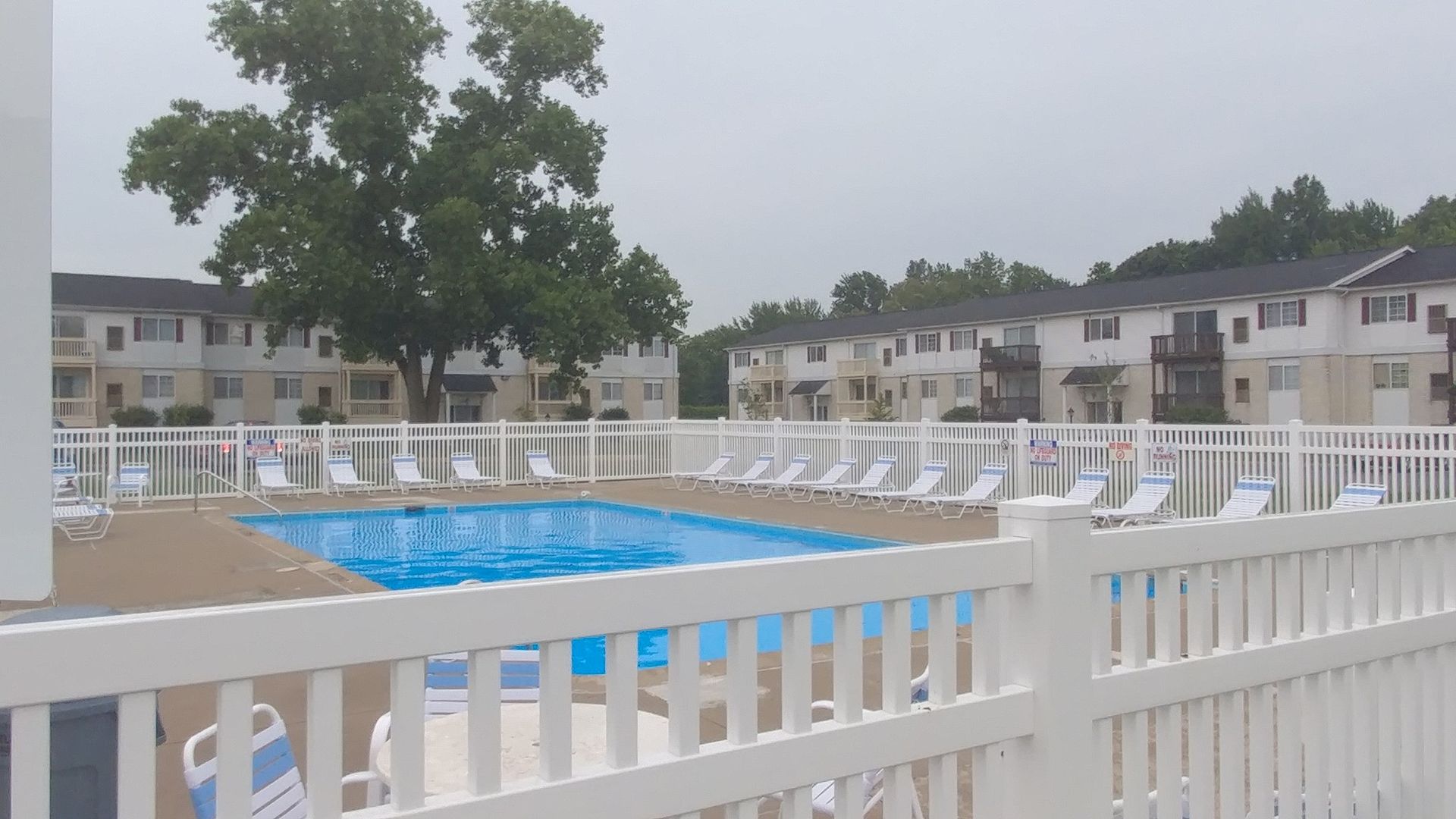 Apartment pool with blue water, white fence, lounge chairs, and trees under an overcast sky