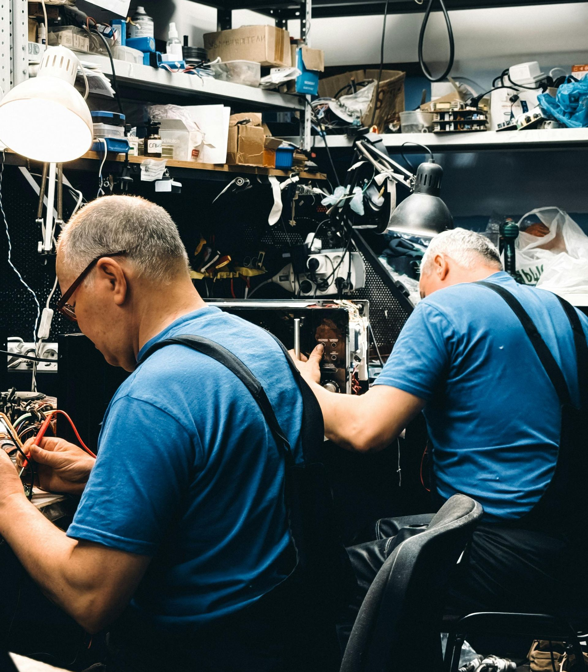 Two people in blue shirts working on electronics in a cluttered workshop.