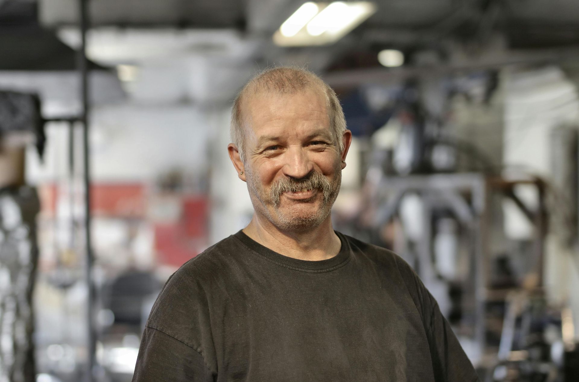 Man with a mustache smiles in a workshop, wearing a dark shirt.