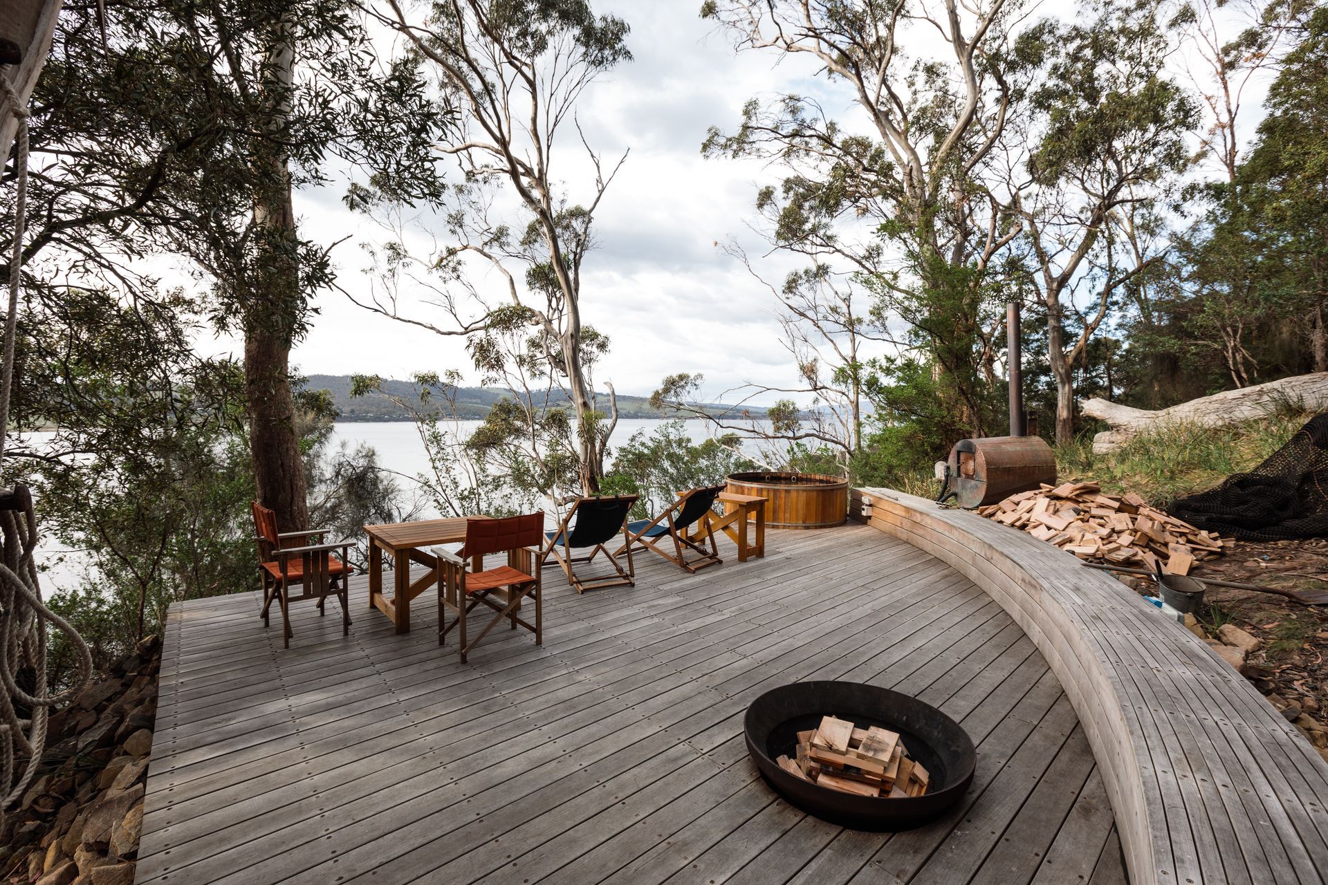 Wooden deck with outdoor dining table, fire pit, and trees overlooking water