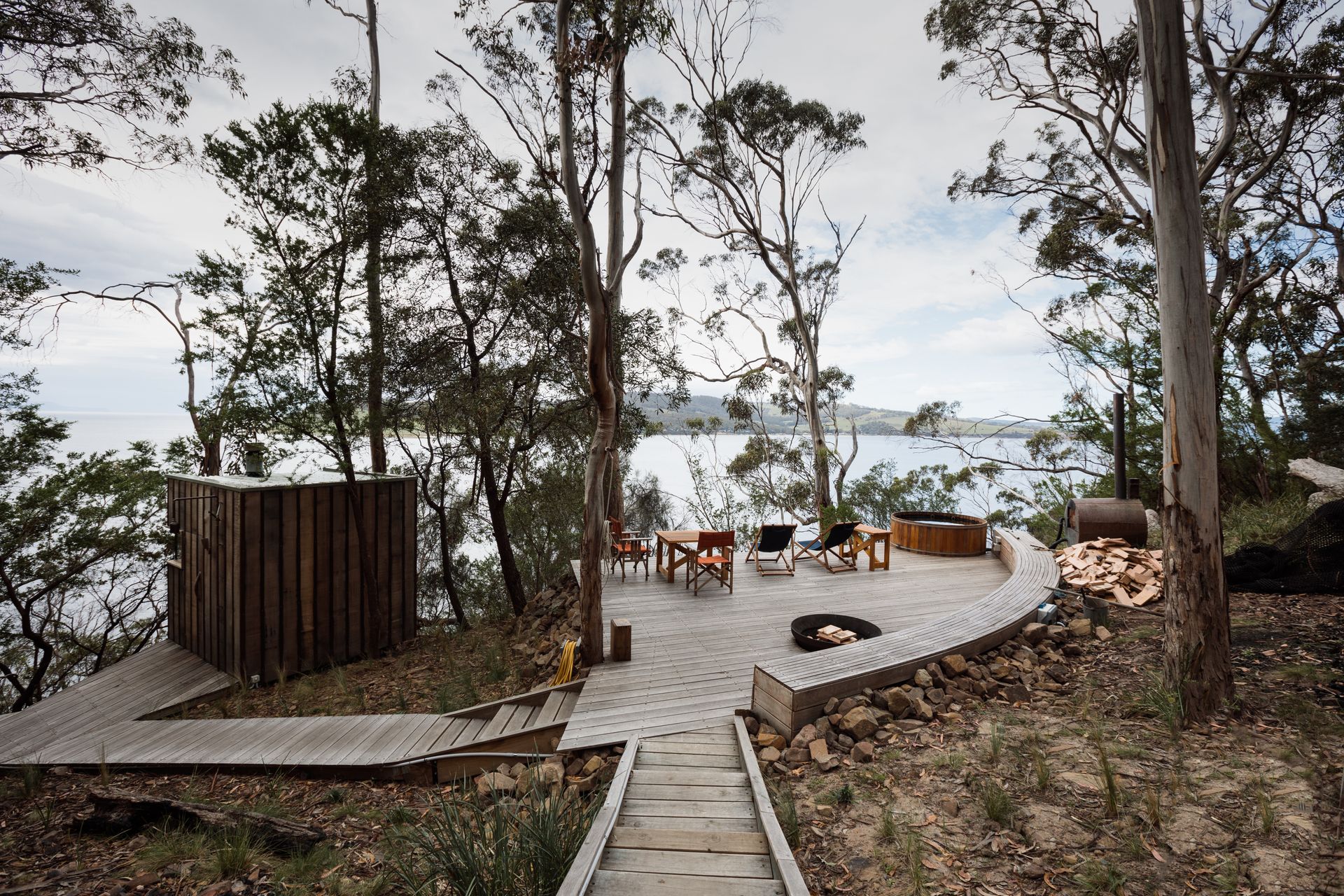 Wooden deck overlooking a lake through trees, with outdoor chairs and a fire pit at dusk