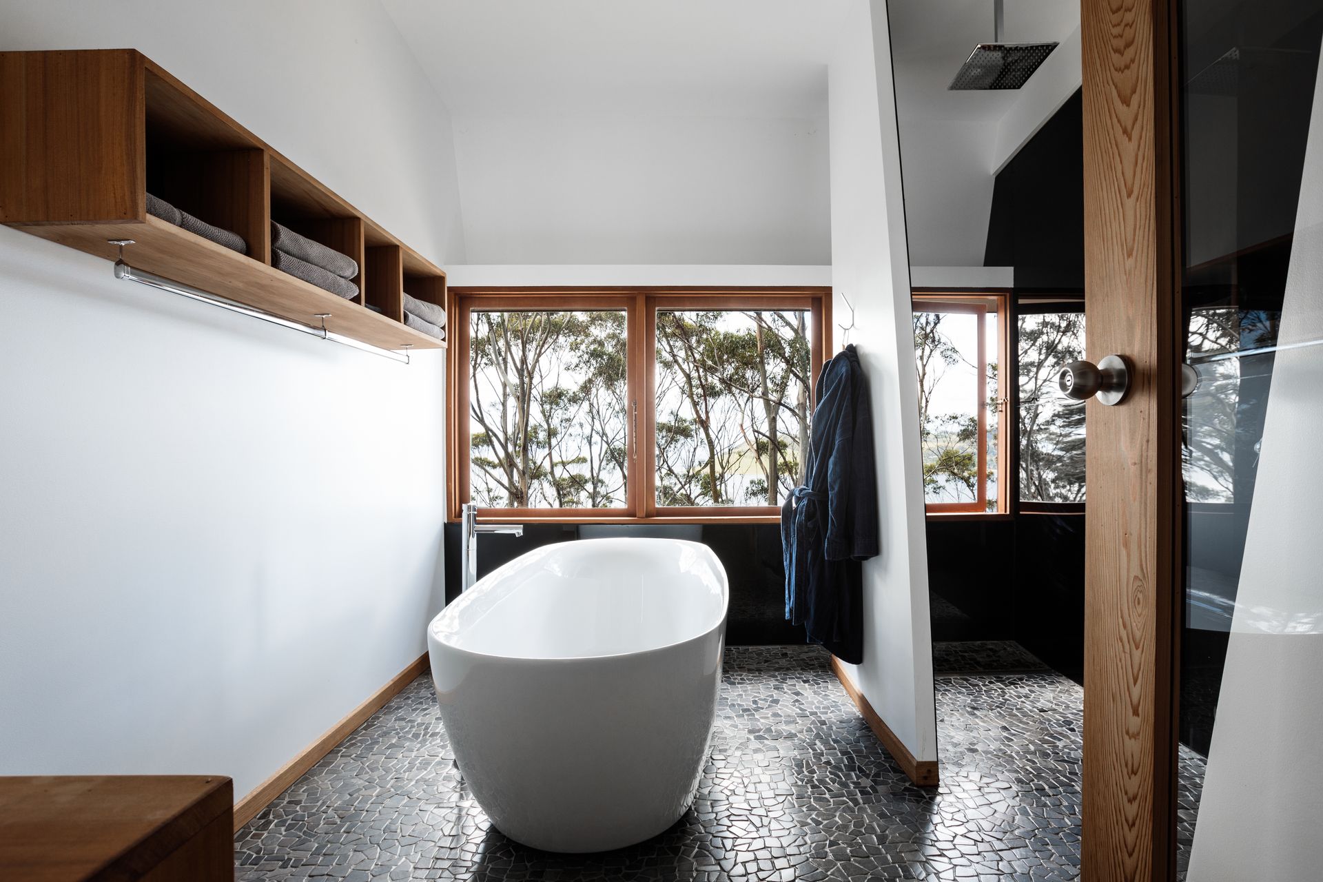 Modern bathroom with a white freestanding tub, wood shelving, and large windows overlooking trees