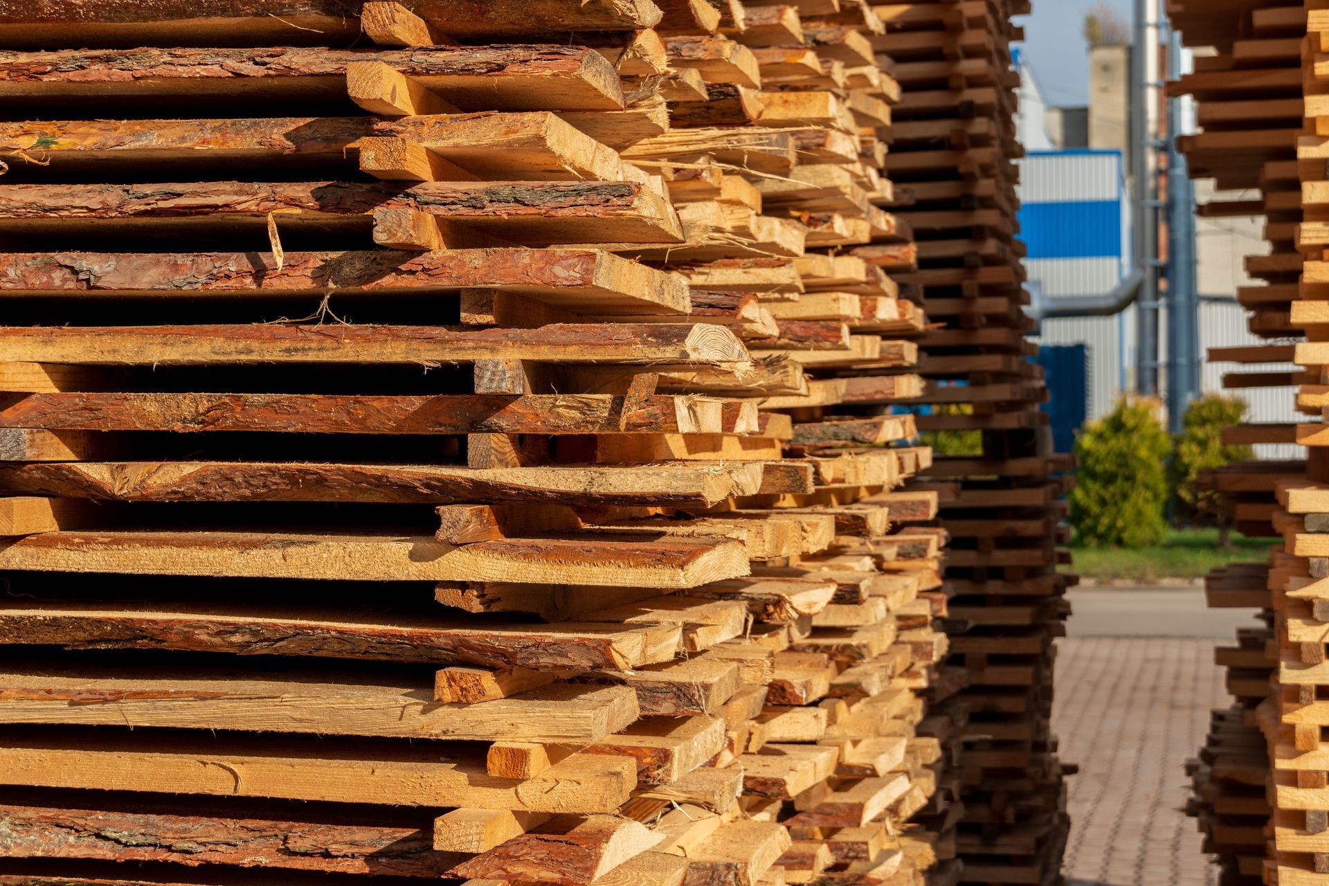 Stacked rough-cut wooden boards piled outdoors beside an industrial building.